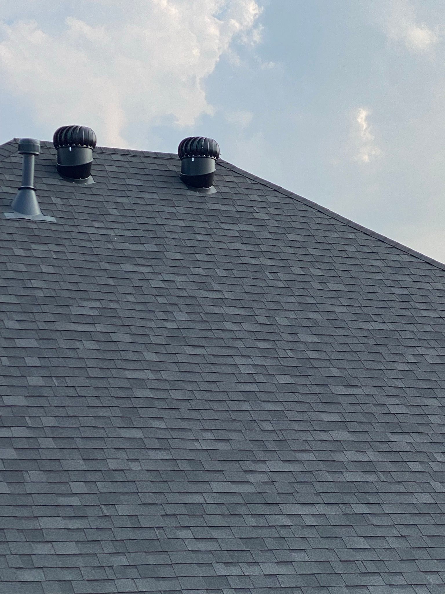 Gray shingle roof with two black turbine vents and a metal pipe against a cloudy sky.