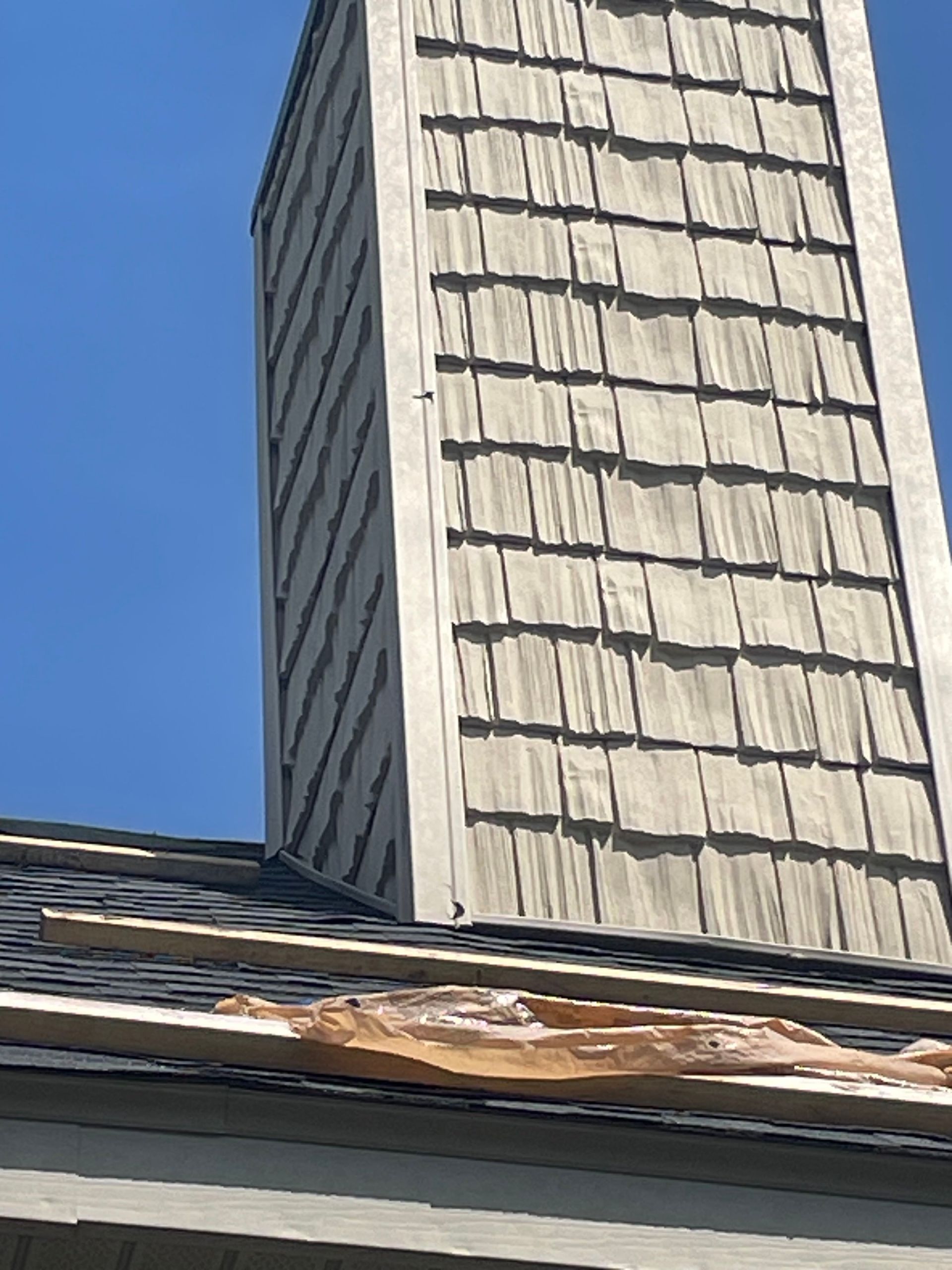 Chimney with weathered shake siding on roof with exposed underlayment against a blue sky.