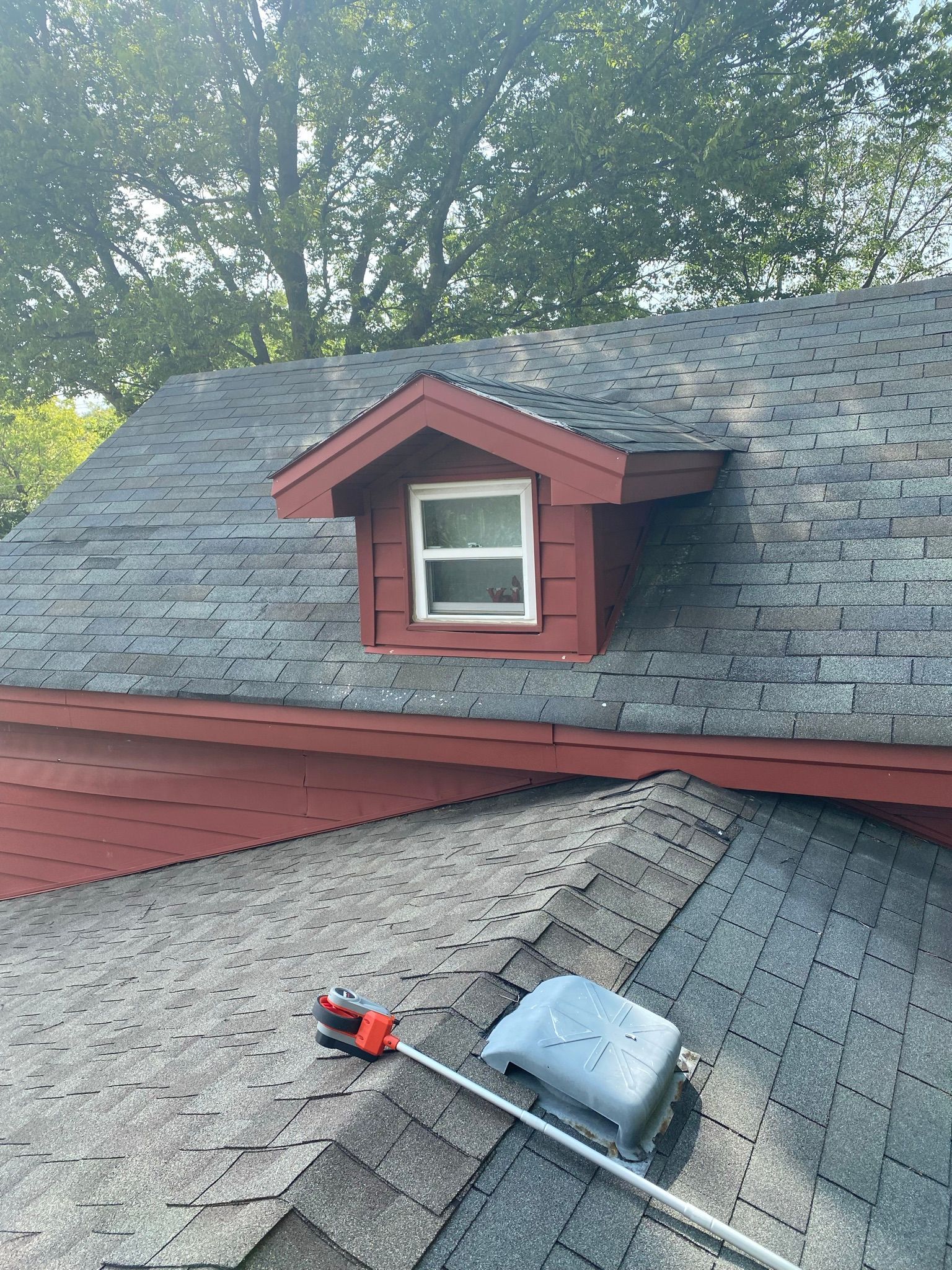 Red dormer with white window on a gray shingled roof, tool on lower roof.