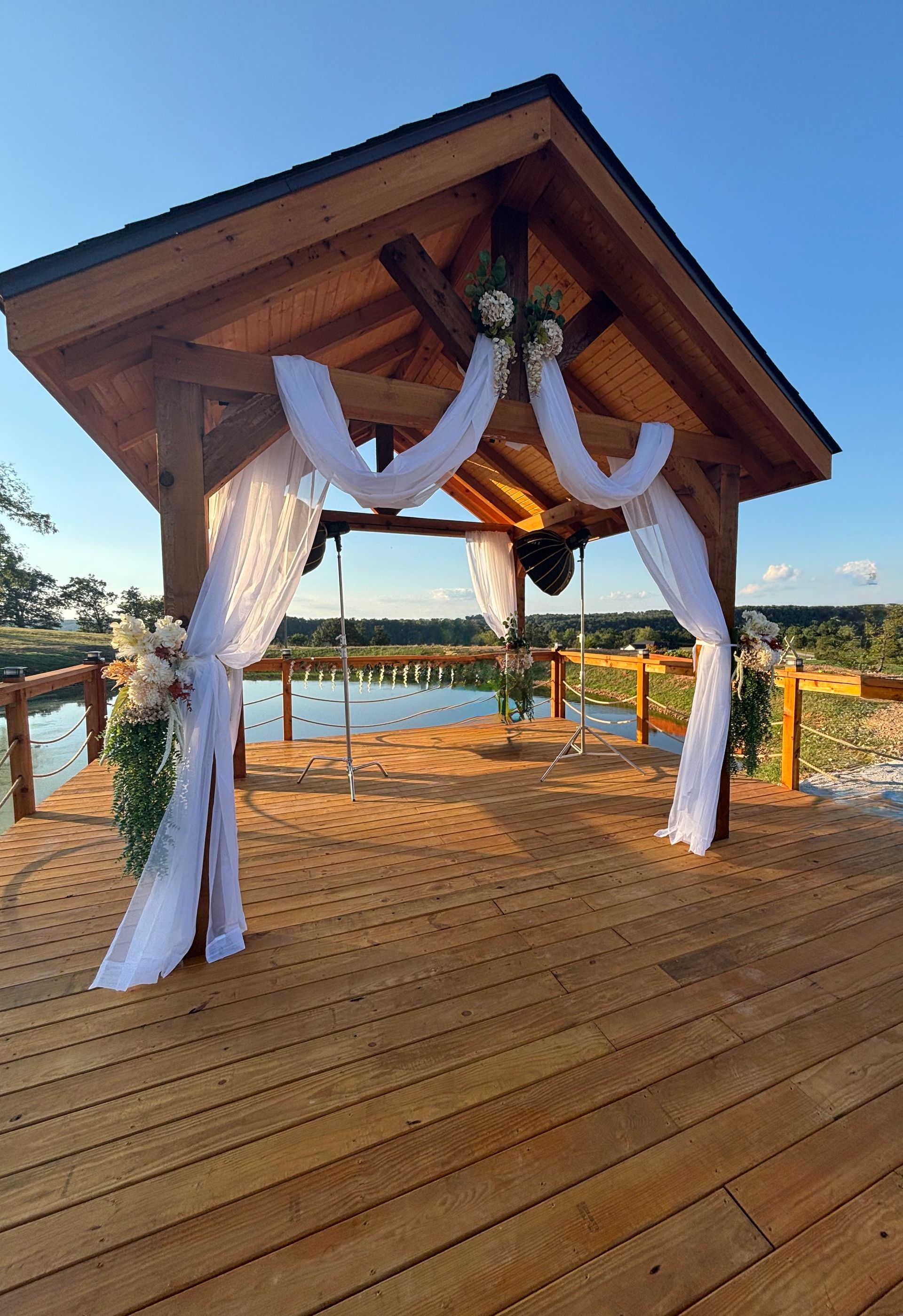 Wooden gazebo decorated for a ceremony overlooking a lake; white fabric drapes, floral accents, clear blue sky.