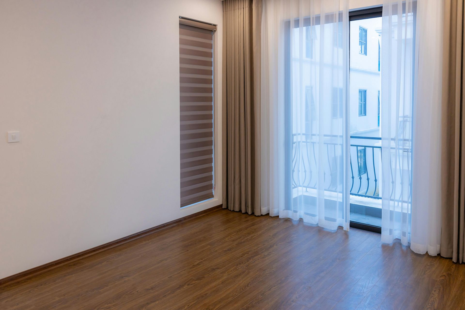 Empty room with wood floors, large window with sheer curtains, and a roller shade.