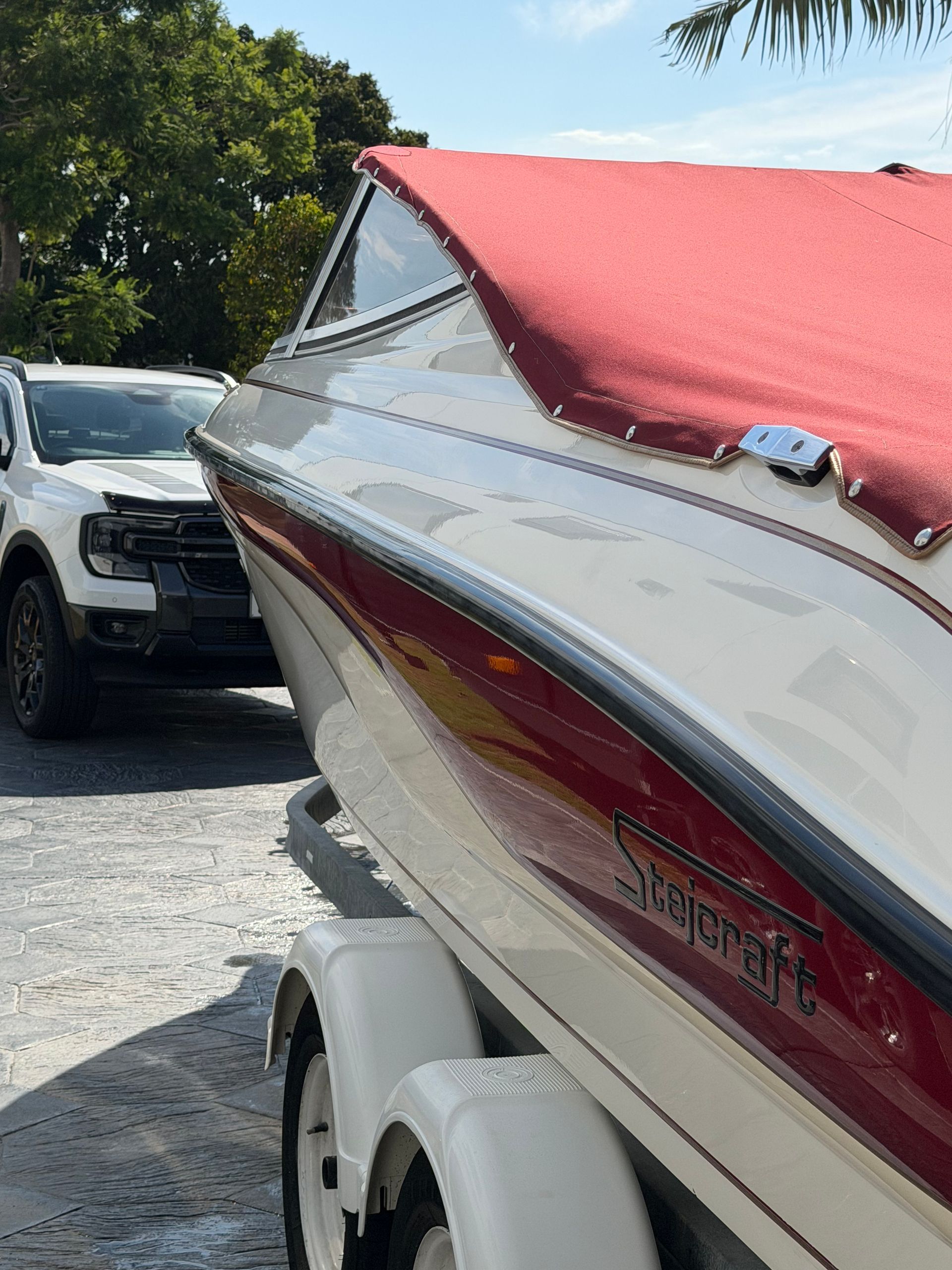 A white and maroon boat on a trailer parked beside a white truck on a gravel lot.