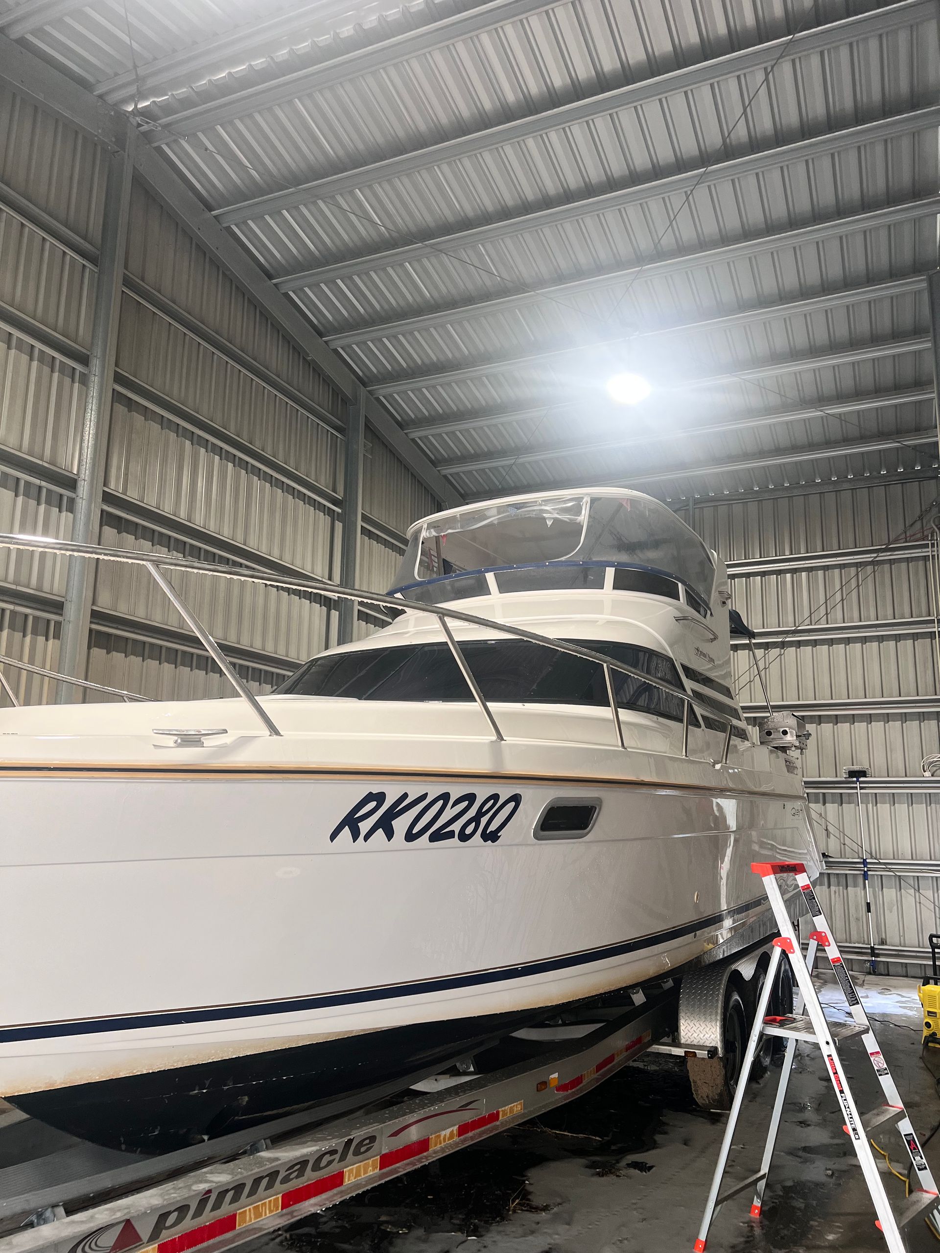 A beige cabin cruiser boat sits on a trailer inside a large metal warehouse, with a ladder standing nearby.