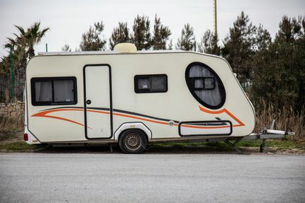 A cream-colored travel trailer with orange and black graphics parked on a gravel lot in front of trees.