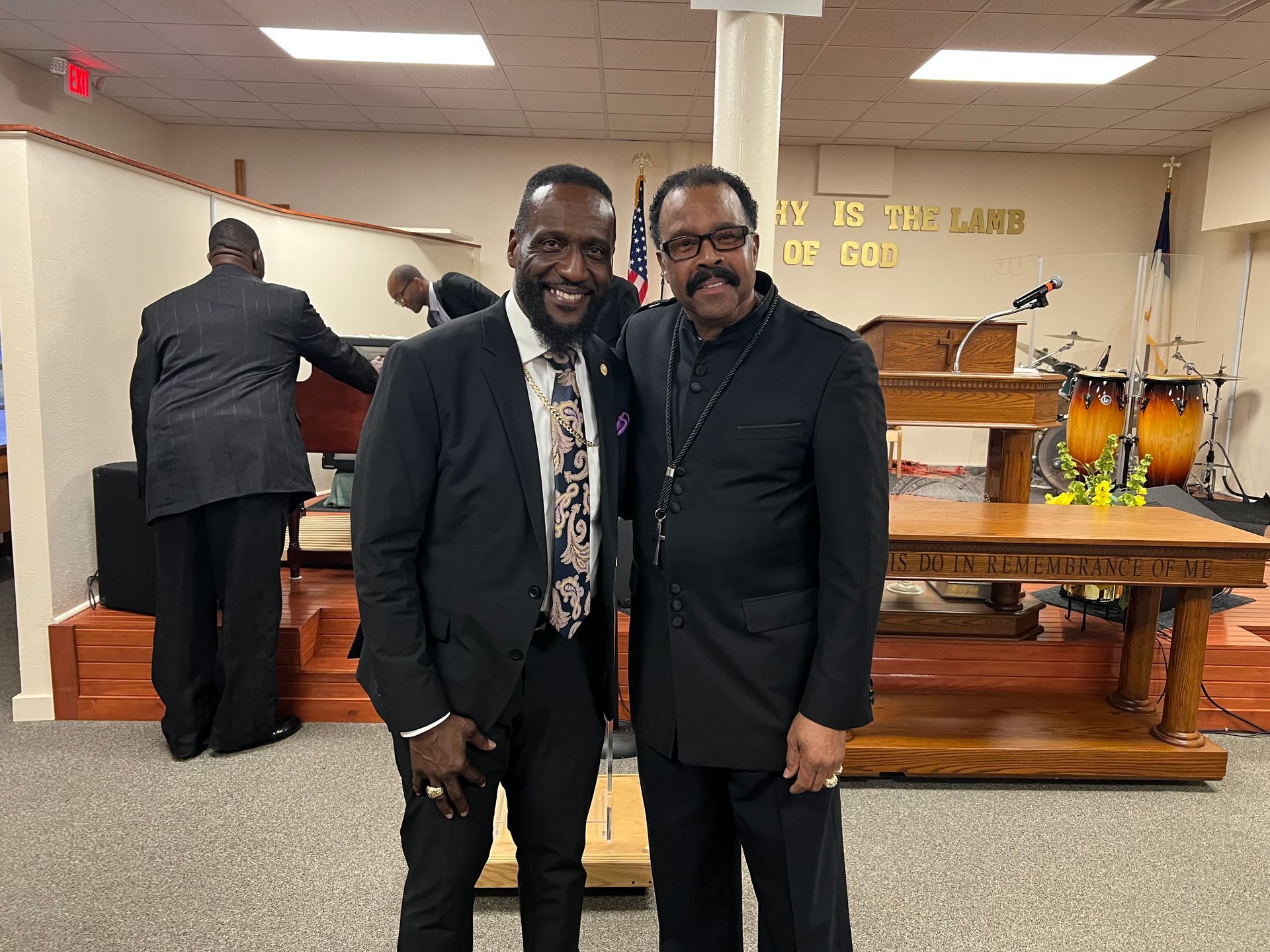 Two men smiling, posing together inside a church. One wears a suit and tie, the other, a black outfit.