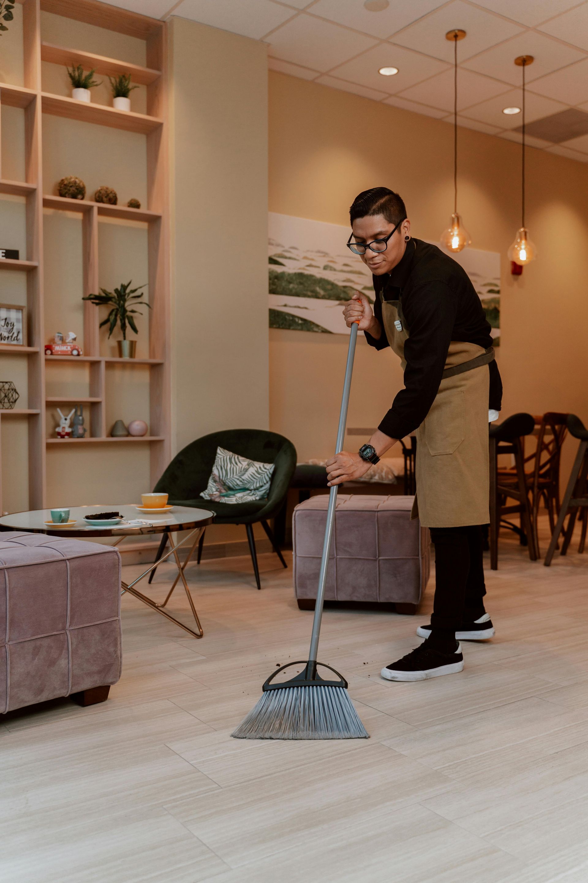 Person sweeping a light-colored floor in a cafe. They wear glasses, a black shirt, and an apron.