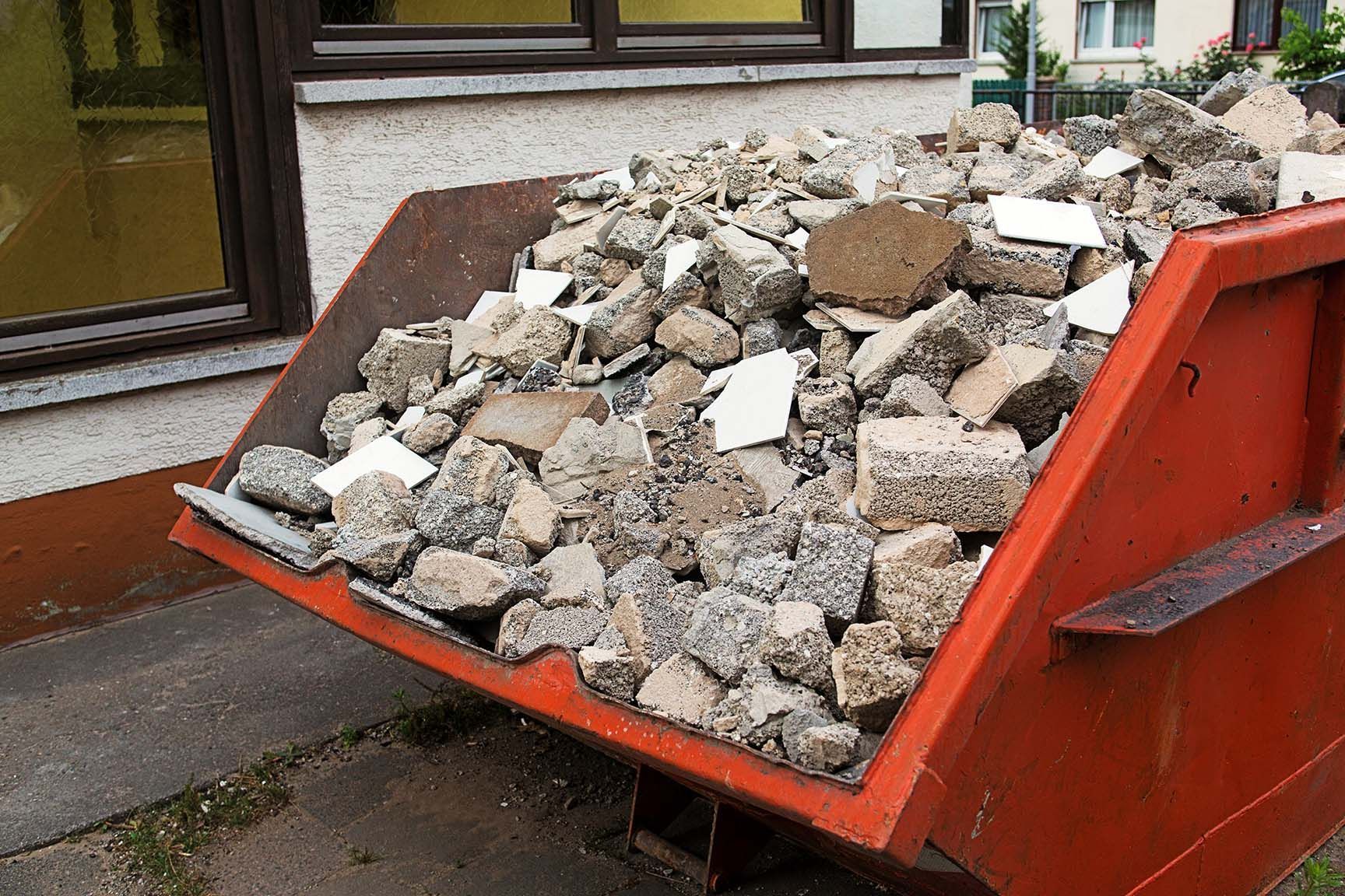 Vor einem Gebäude steht ein mit Steinen gefüllter Müllcontainer.