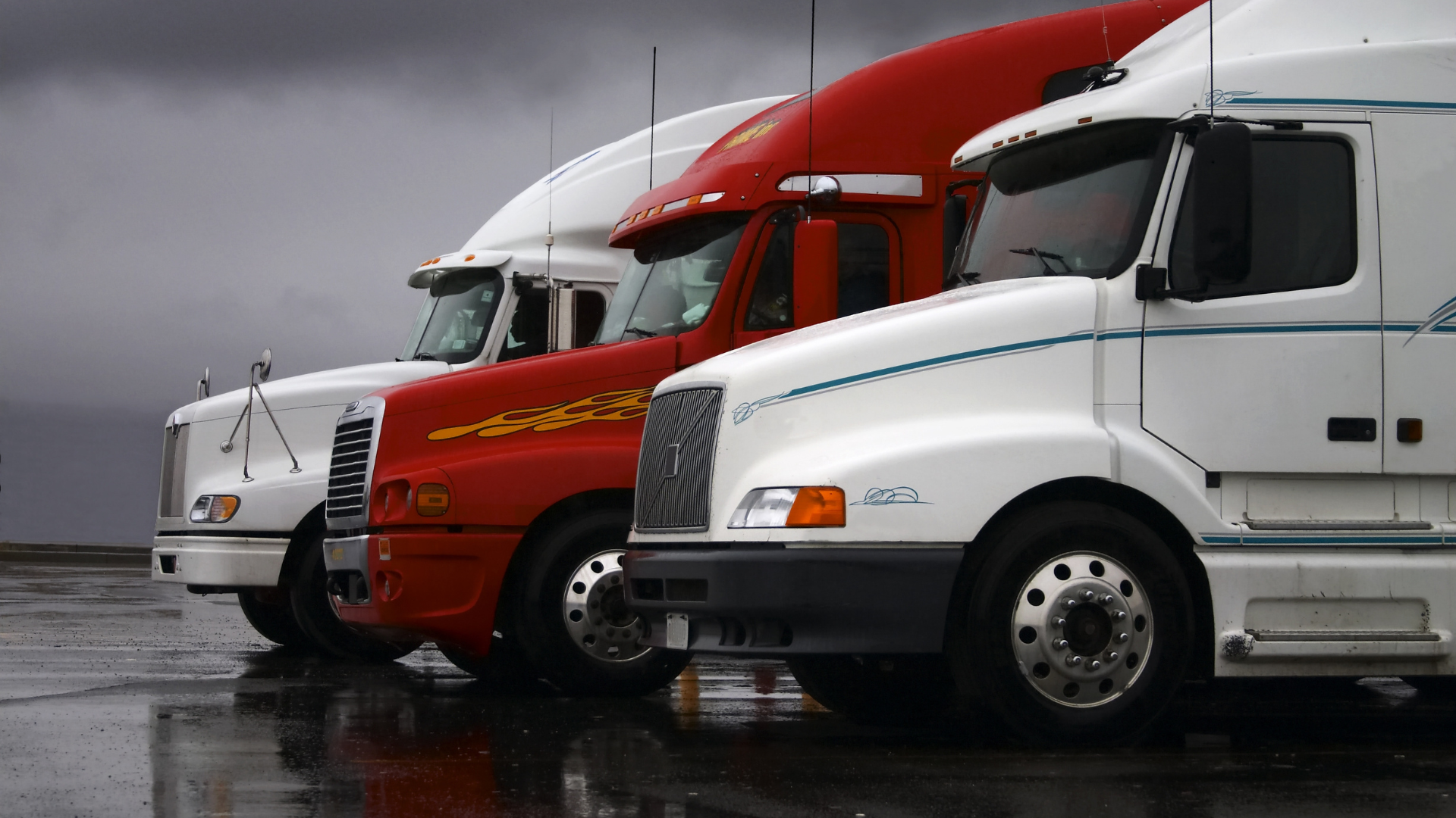 Three semi-trucks parked in a row; white, red, and white, against a gray, rainy sky.