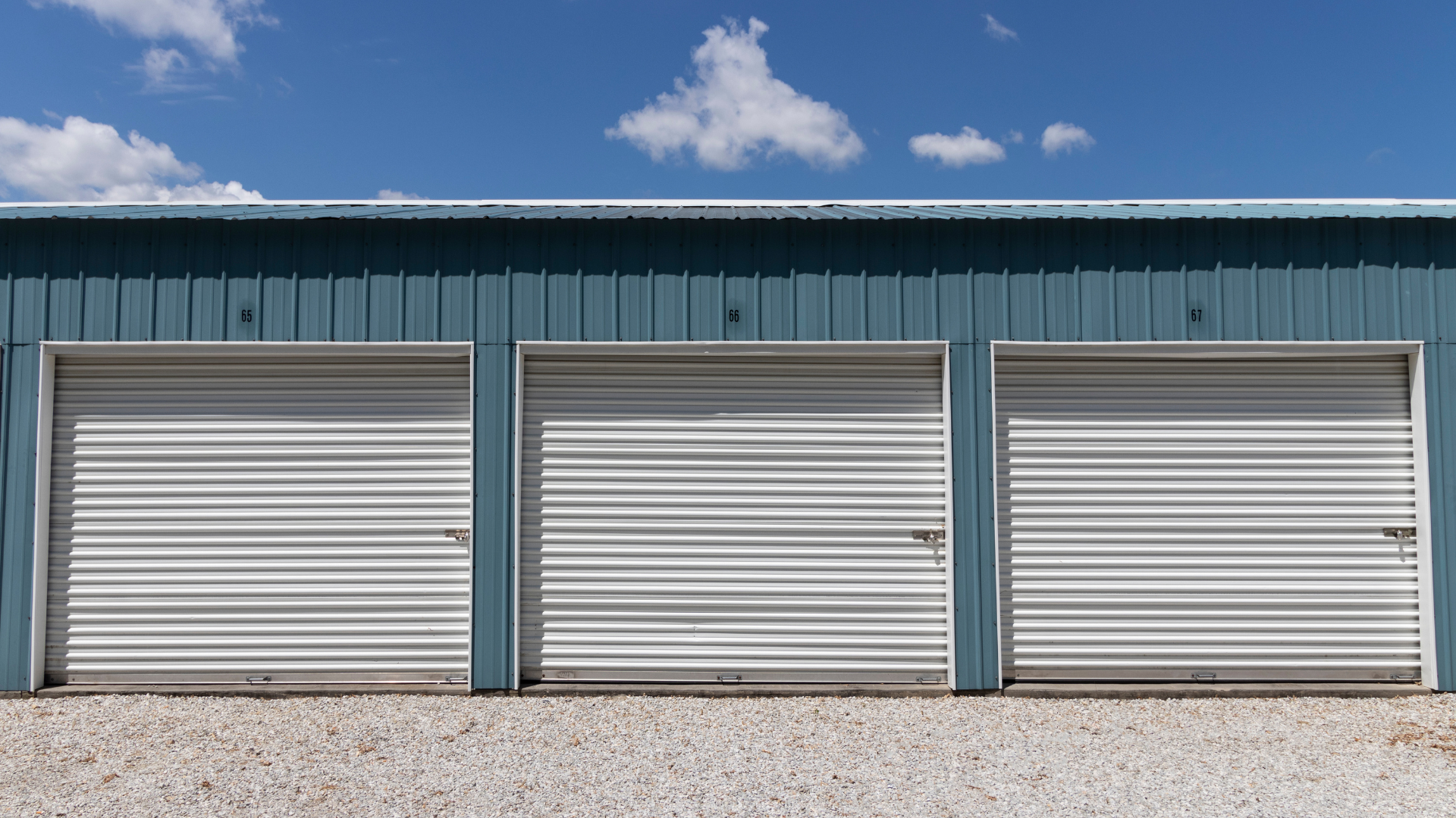 Three closed storage unit doors with white metal shutters. Blue building, gravel ground, blue sky.