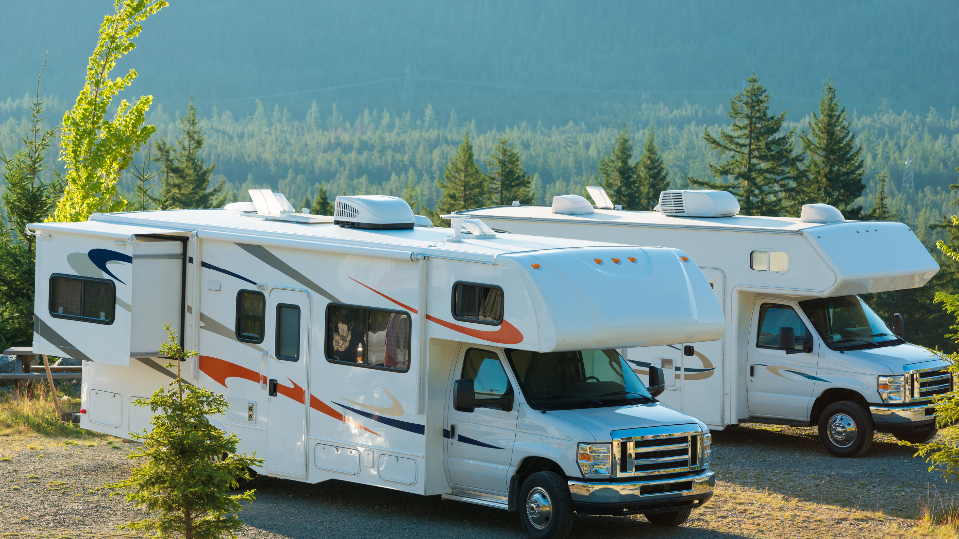 Two white RVs parked on a gravel lot in front of a forested mountain. People are visible in the window of one RV.
