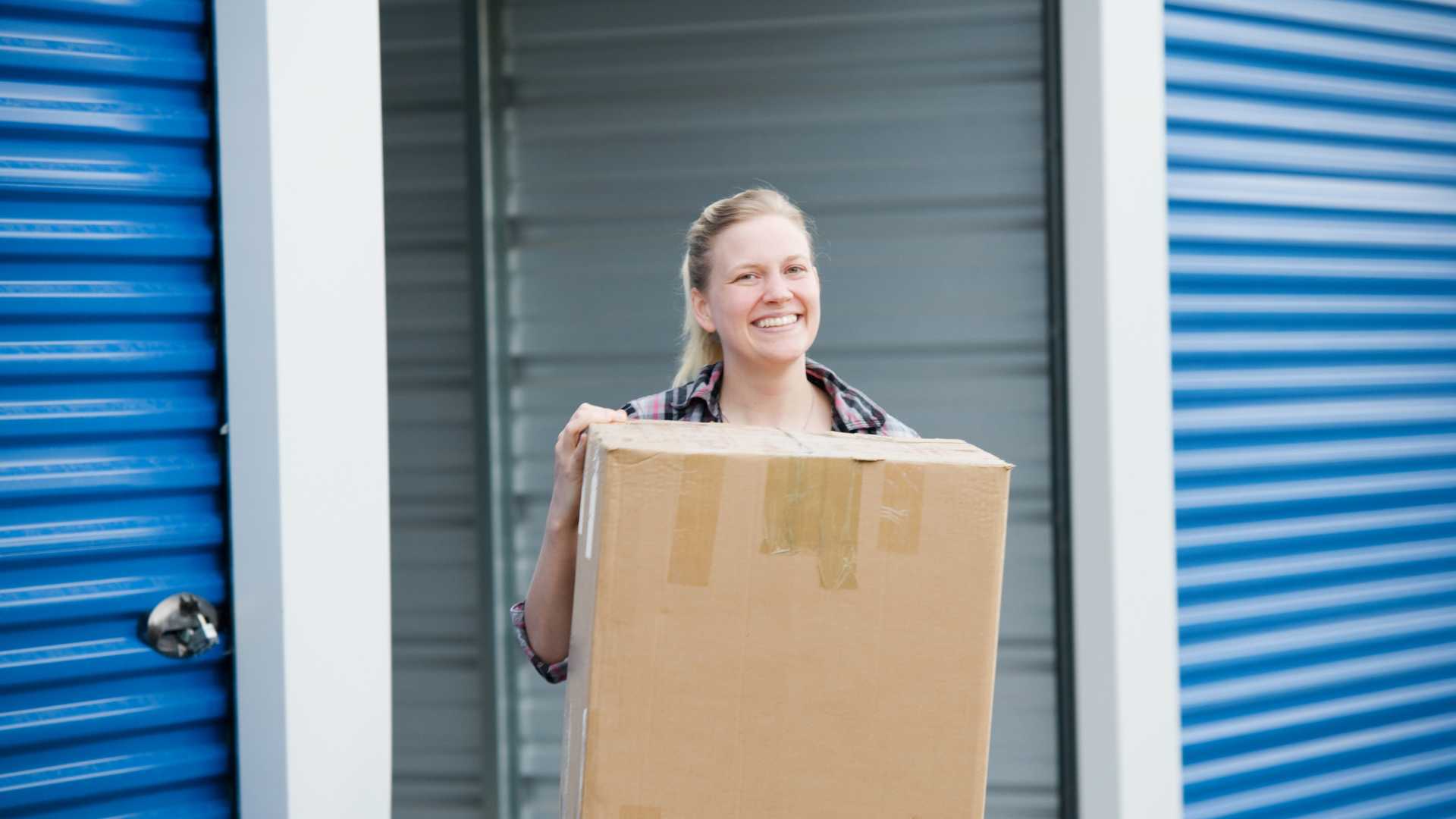 Woman smiling, carrying cardboard box out of blue storage unit.