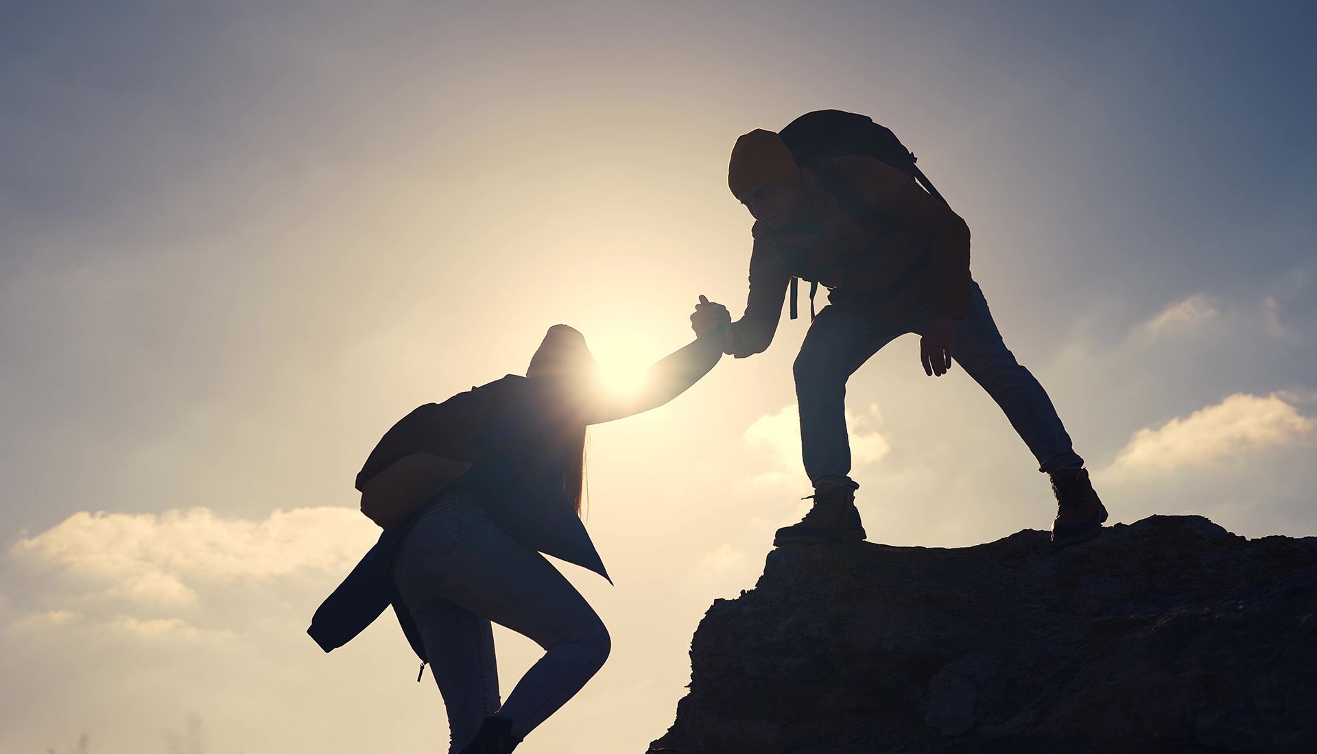 Hikers helping each other climb a rocky peak, silhouetted against the sunlit sky.