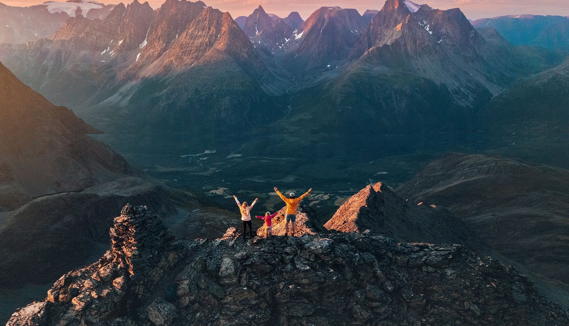 People with arms raised on a rocky mountain summit at sunset, mountain range in background.