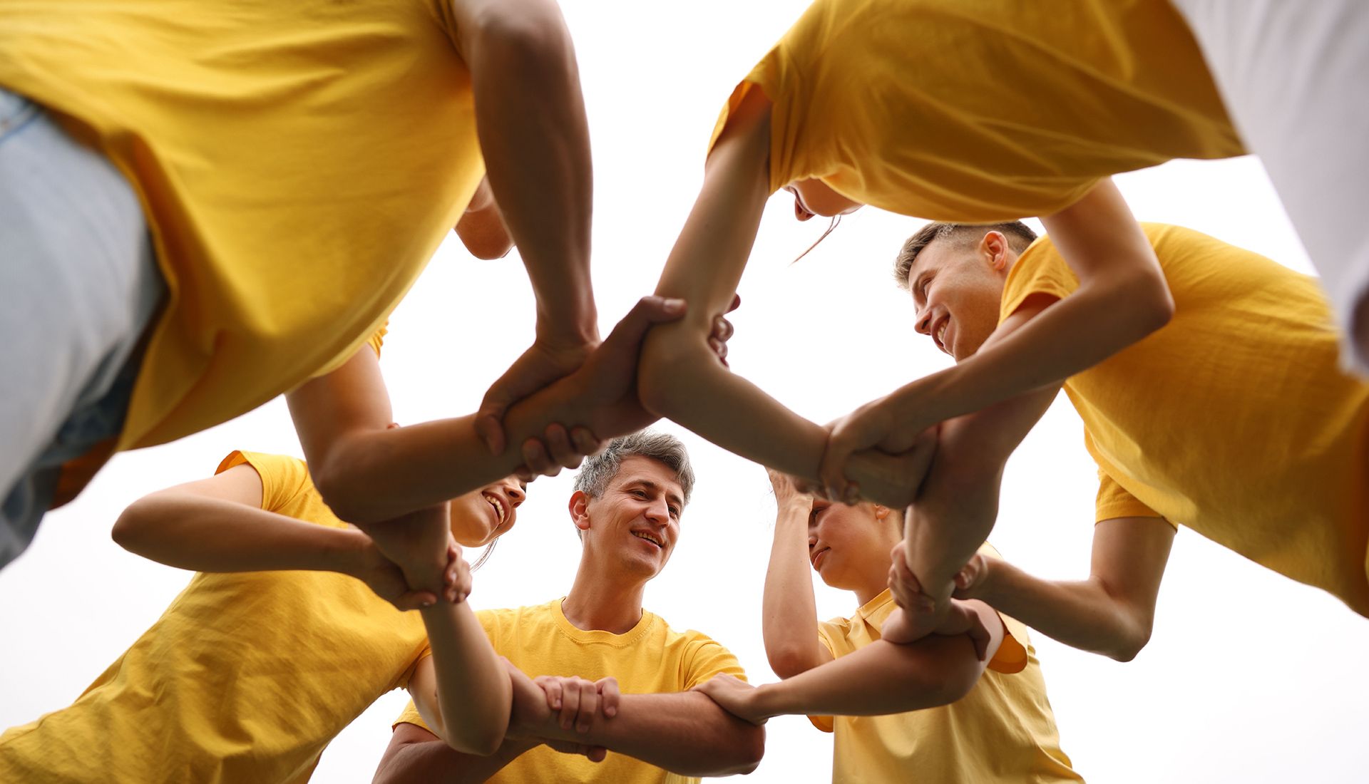 People in yellow shirts forming a circle, holding each other's arms against a white sky.