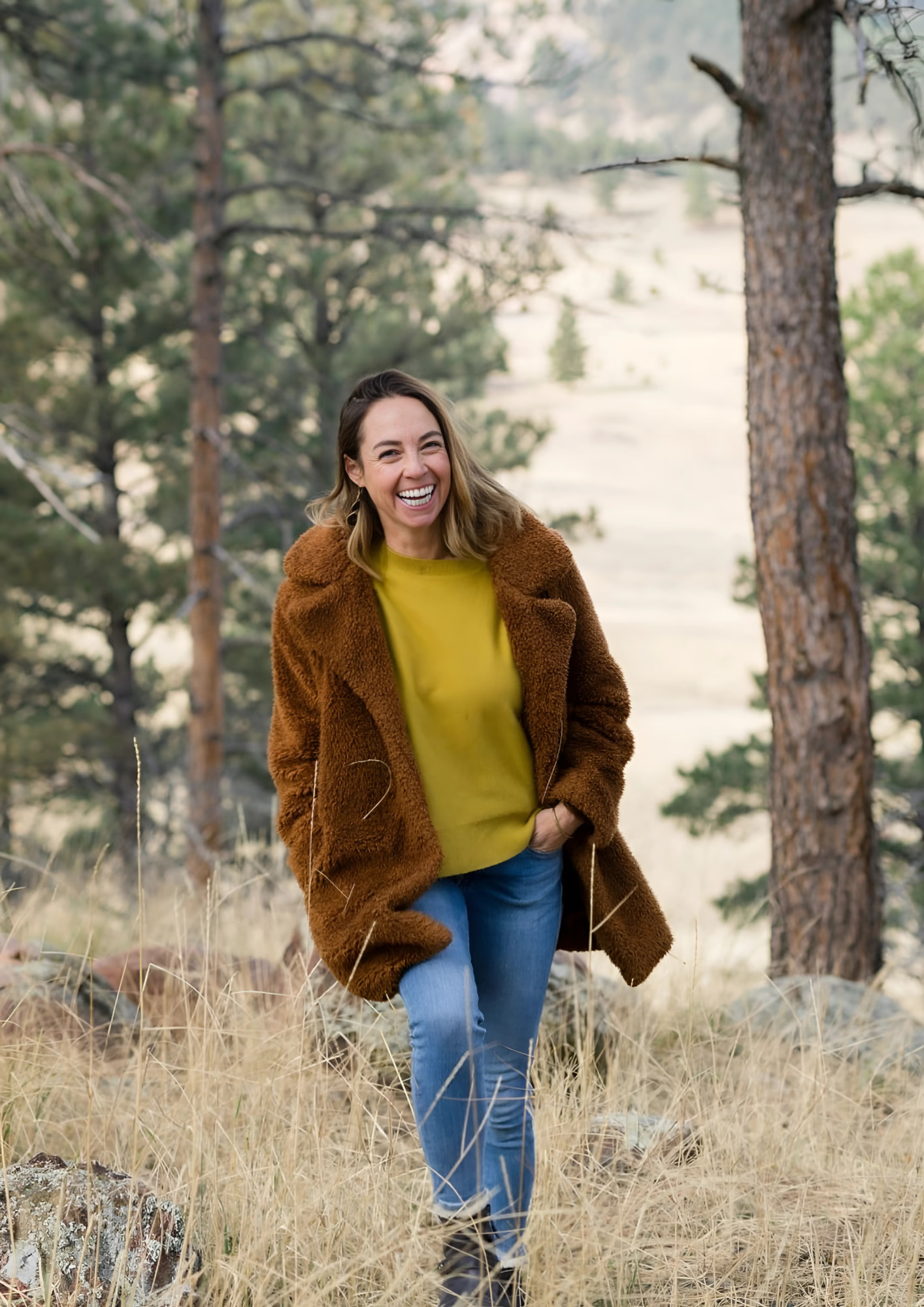 Woman in brown teddy bear coat and yellow sweater smiles outdoors.