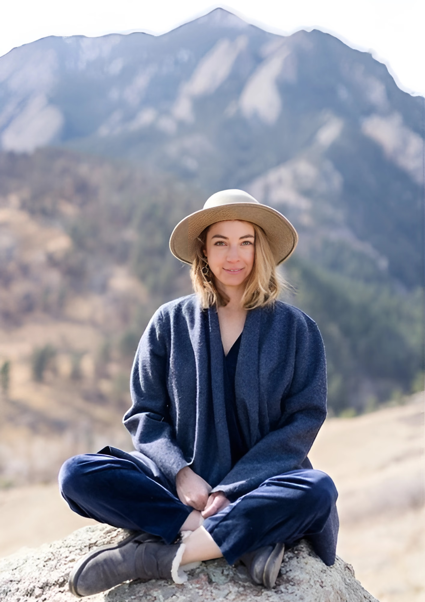 Woman in blue outfit and hat sitting cross-legged on a rock with mountains in the background.