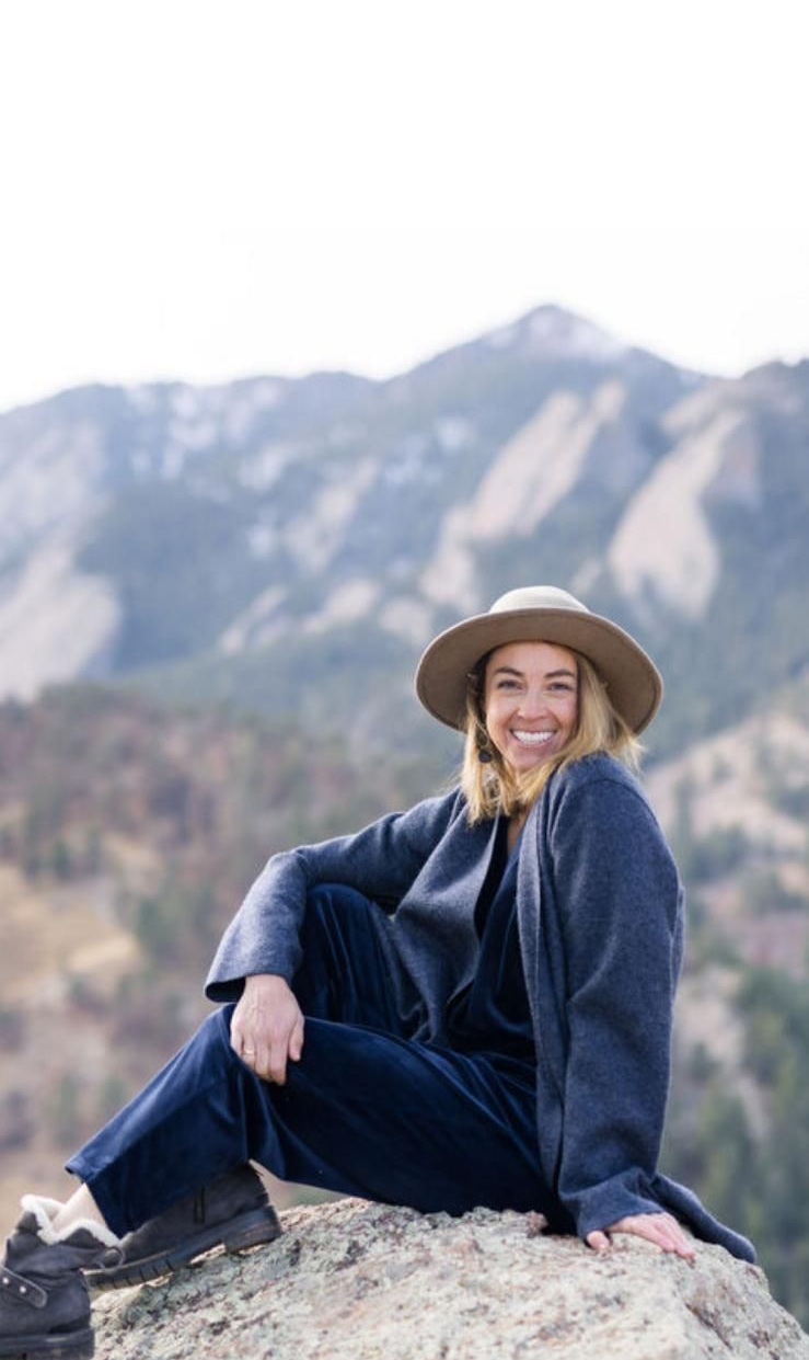 Woman in a brown teddy bear coat and hat smiles outdoors near trees.