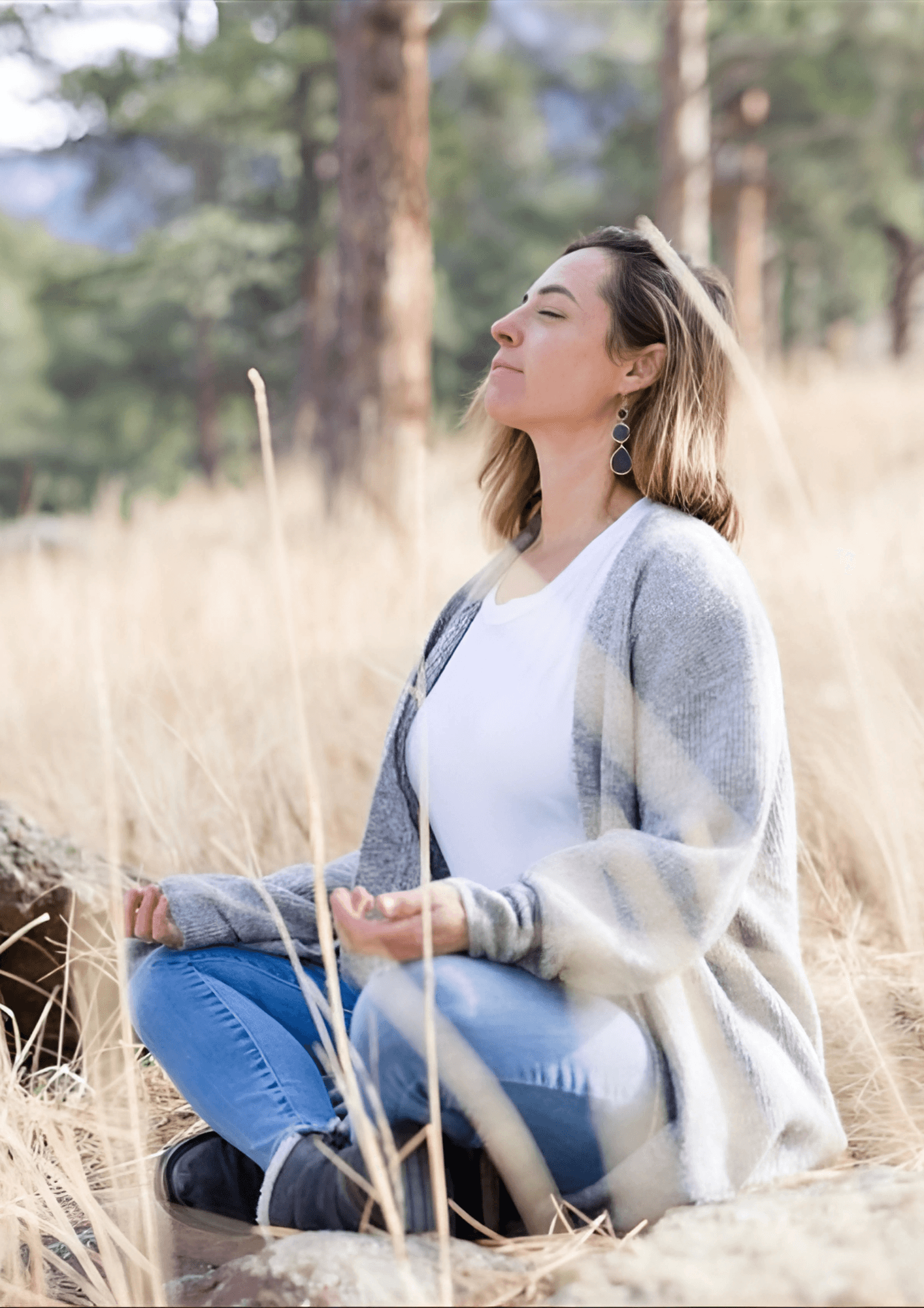 Woman meditating outdoors, eyes closed, hands in lap, gray cardigan, blue jeans, dry grass background.