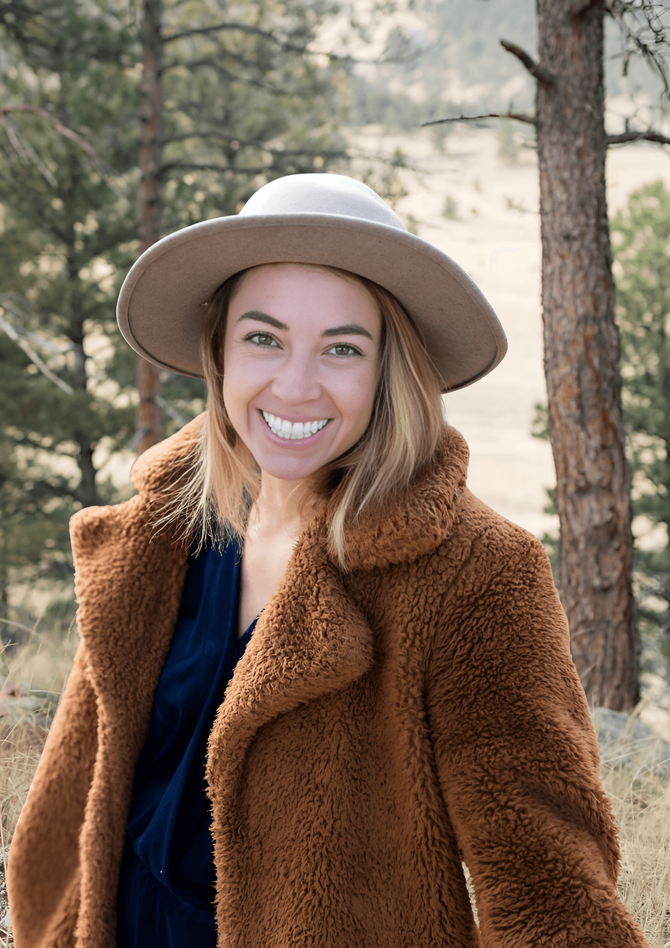 Woman in a brown teddy bear coat and hat smiles outdoors near trees.