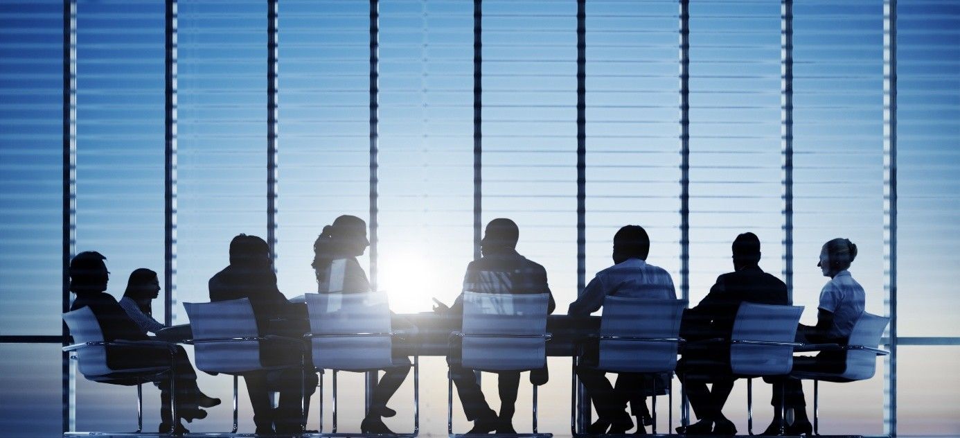 Een zakelijk team zit aan een vergadertafel met grote ramen, silhouetten en fel licht.