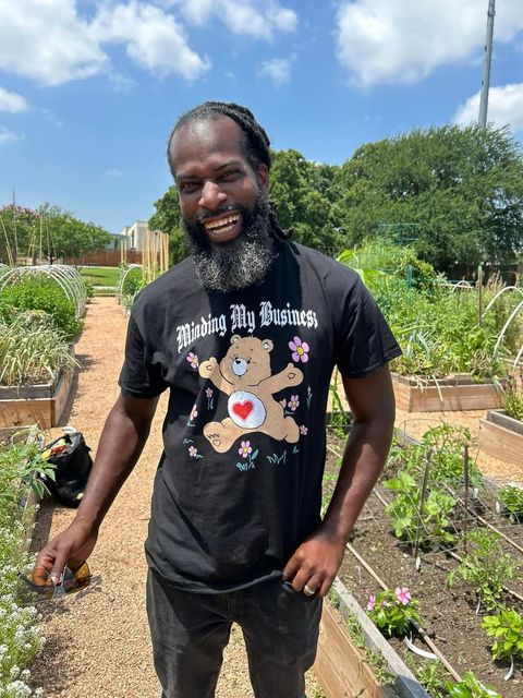 A man with a beard wearing a care bear shirt is standing in a garden.