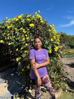 A woman in a purple shirt is standing in front of a bush with yellow flowers.