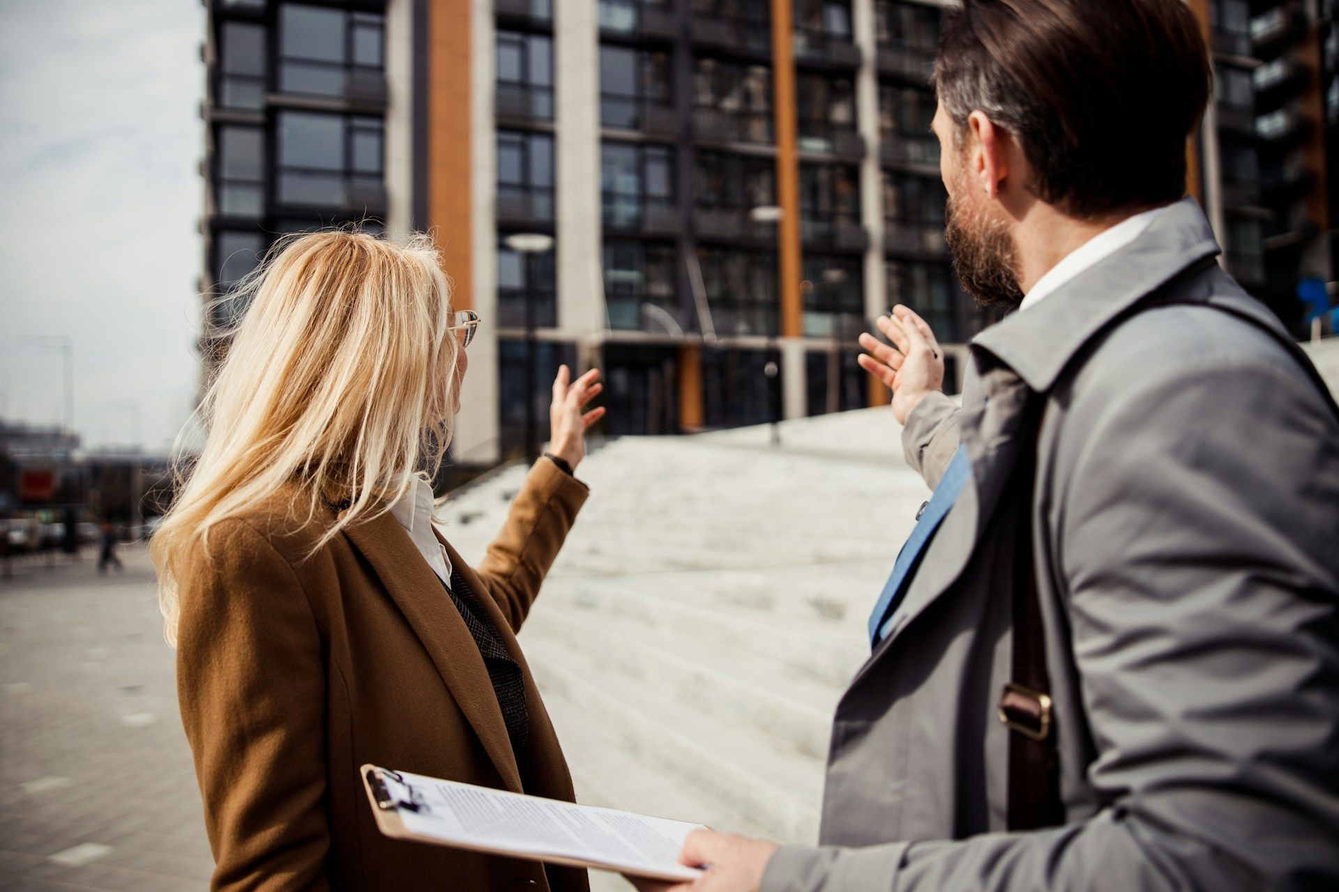 Two people in coats gesture toward a modern building under construction.
