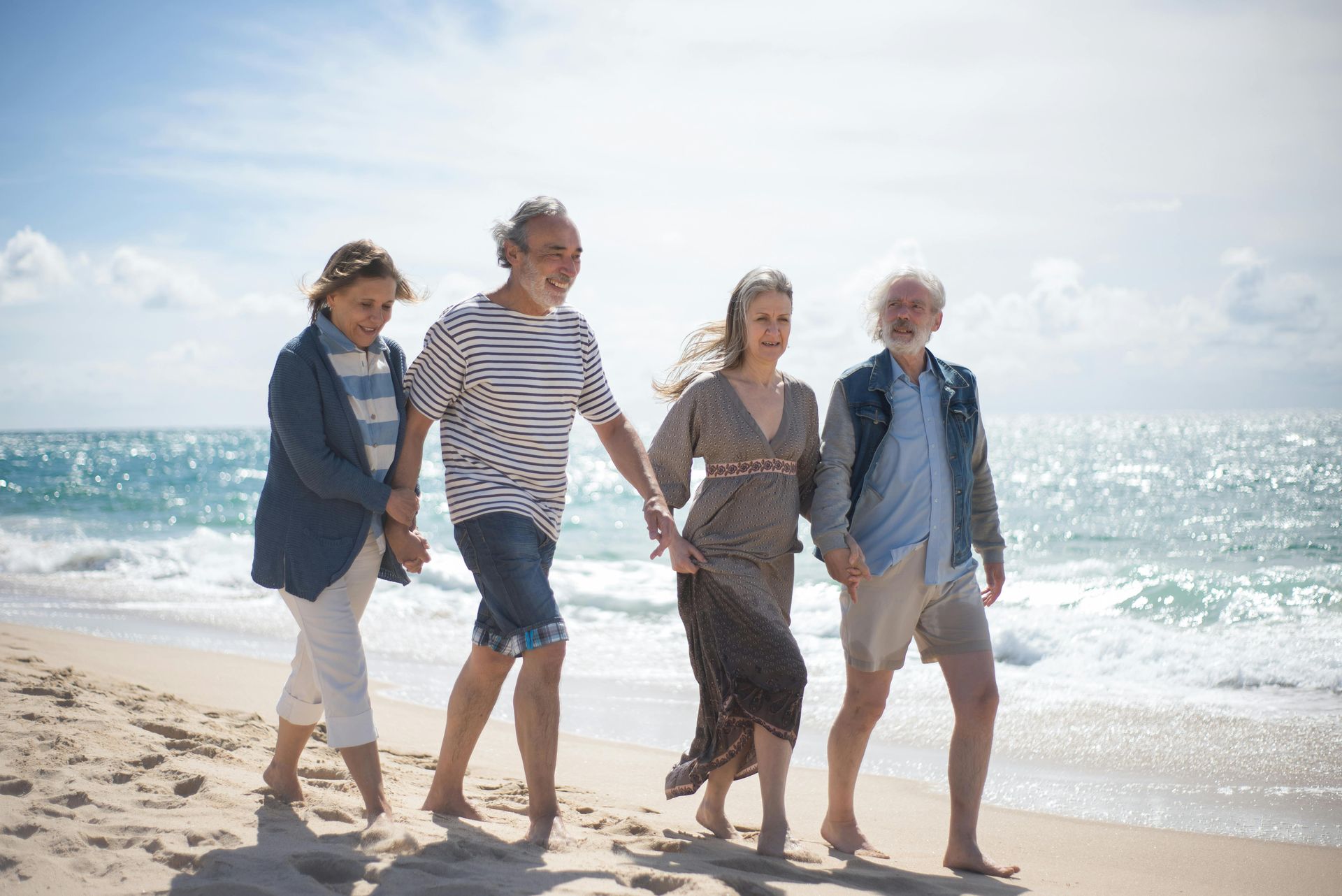 Four friends walking barefoot along a sunny beach by the ocean.