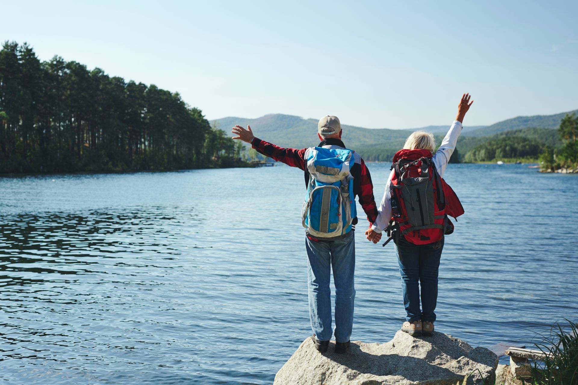 A man and a woman are standing on a rock near a lake with their arms in the air.