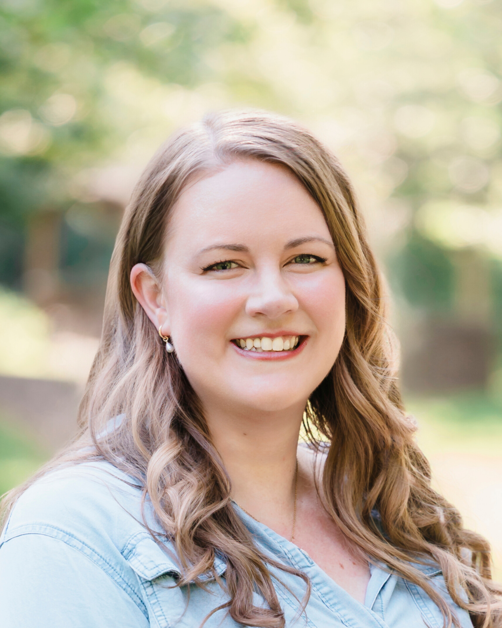 Woman with long wavy hair smiles outdoors in a denim shirt.