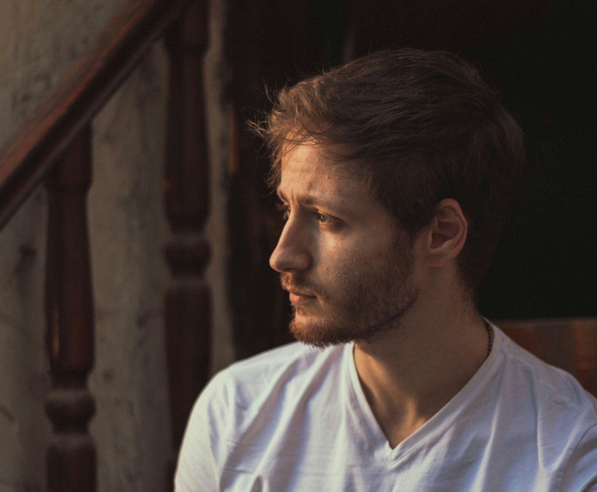 Man with brown hair pulled back, wearing a white t-shirt, looking off to the side. Natural light.