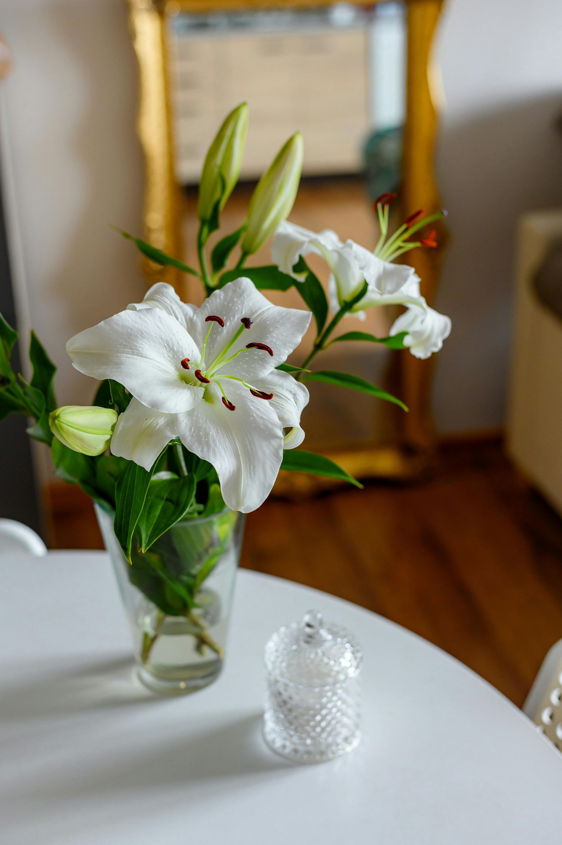 White lilies in a vase on a white table, a clear glass thimble, and a gold-framed mirror in the background.