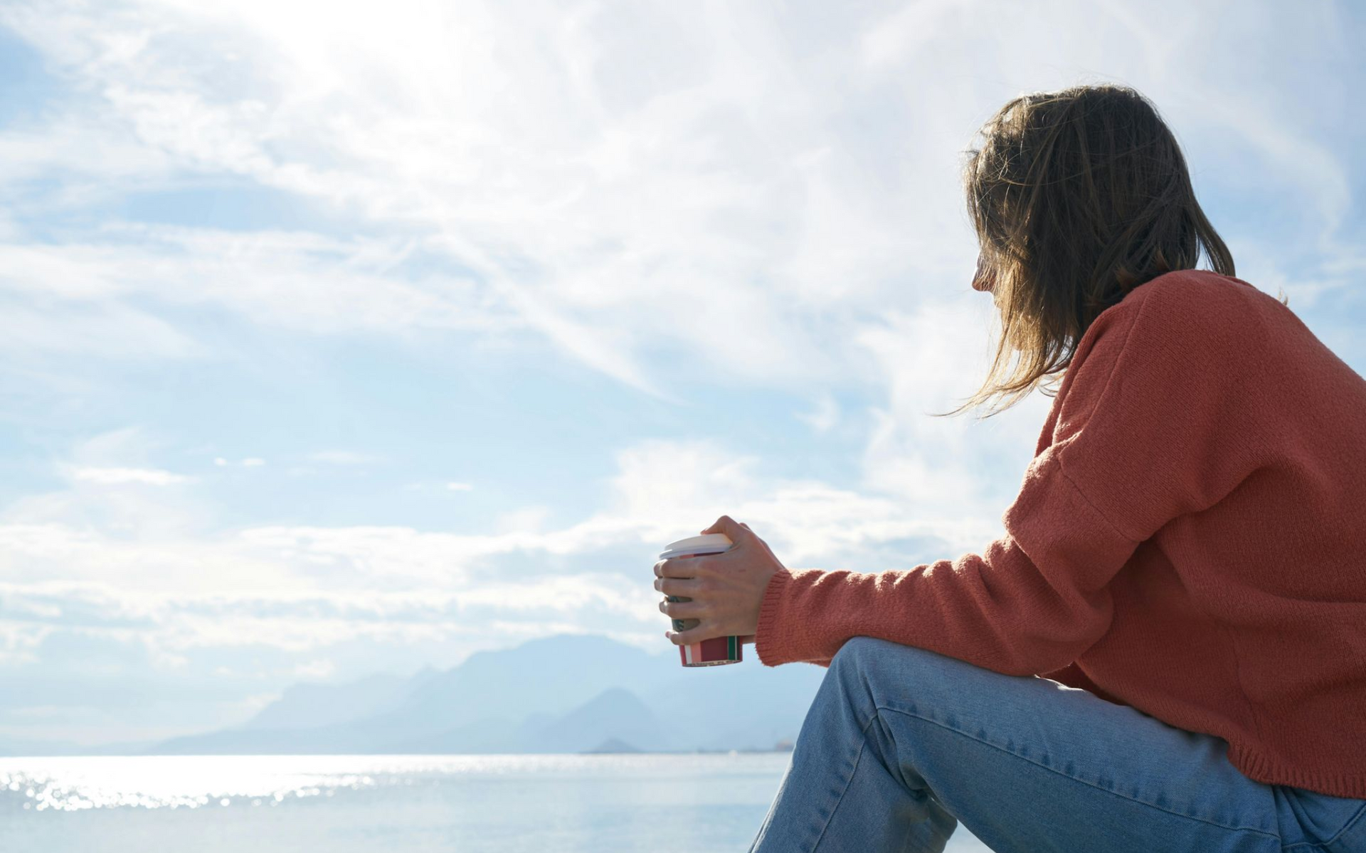 Woman in orange sweater and jeans holds a cup, looking at a seascape with mountains under a sunny sky.