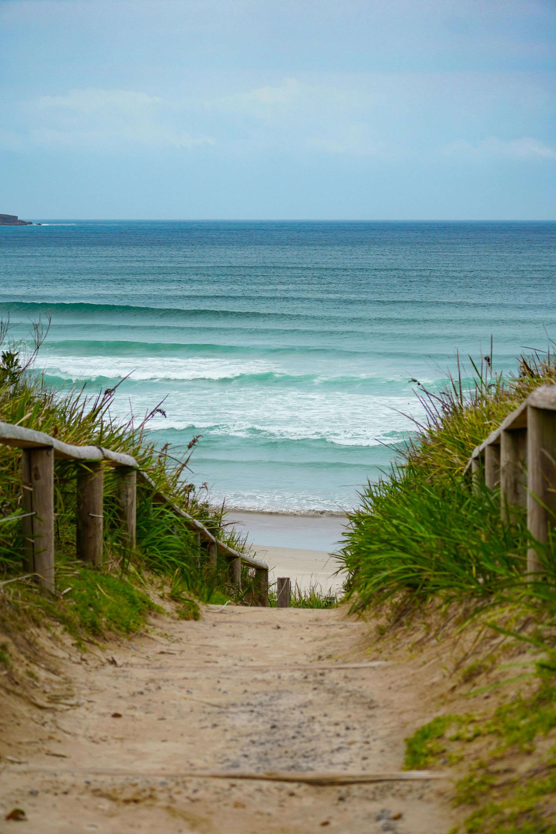 Sandy path with wooden railings leads to a beach with ocean waves under a cloudy sky.