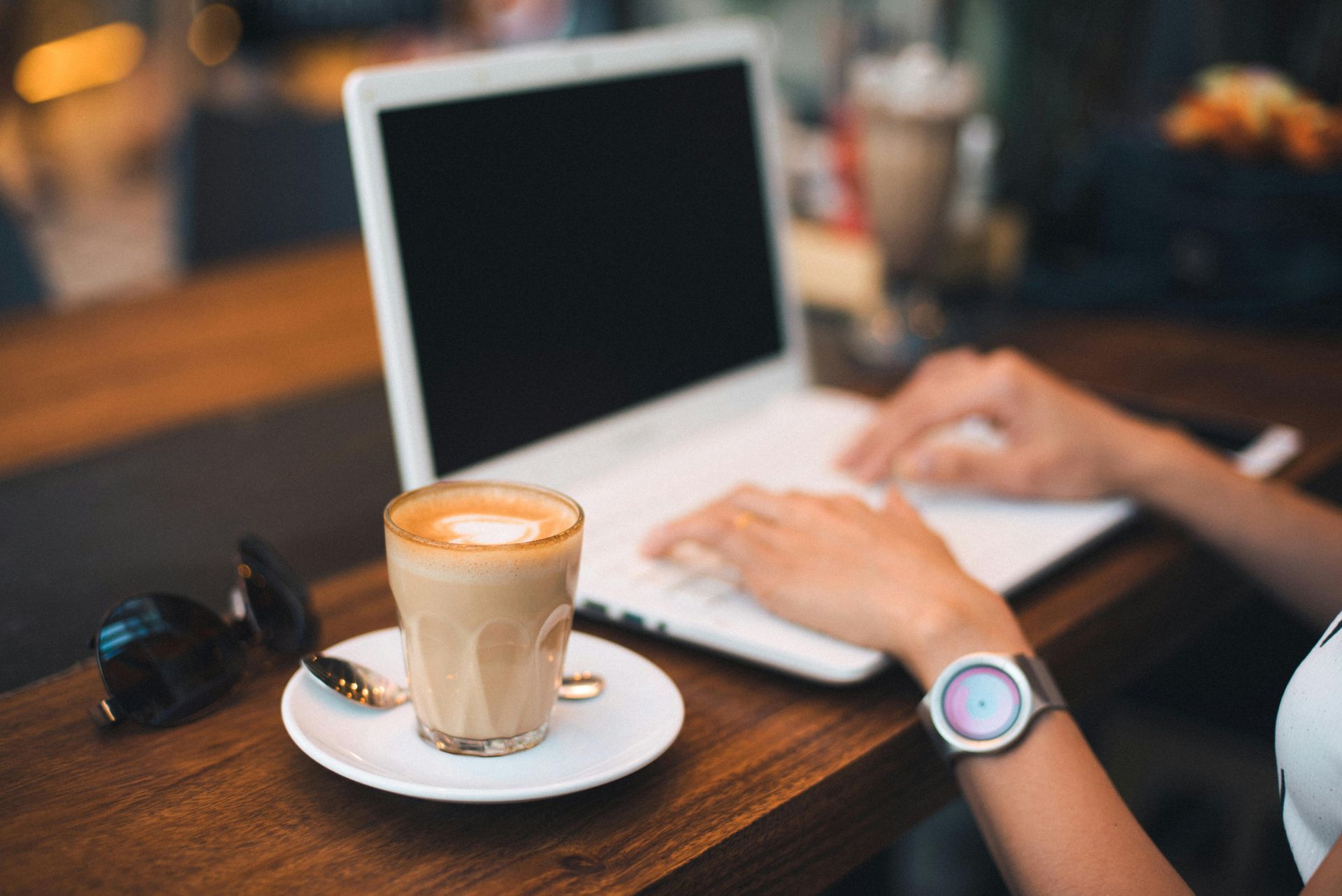 Person typing on a laptop at a cafe with a latte and sunglasses.