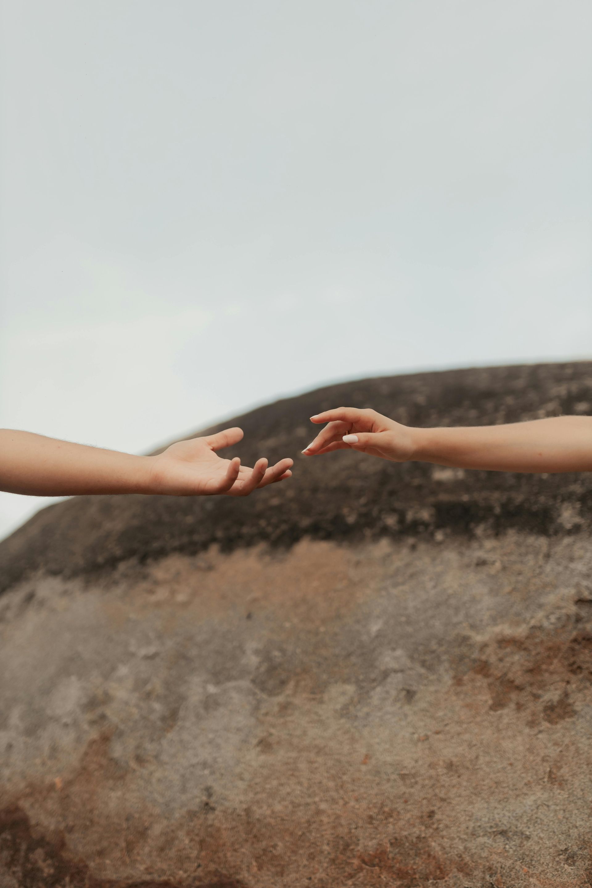 Two hands reaching towards each other, against a blurred rock and sky background.