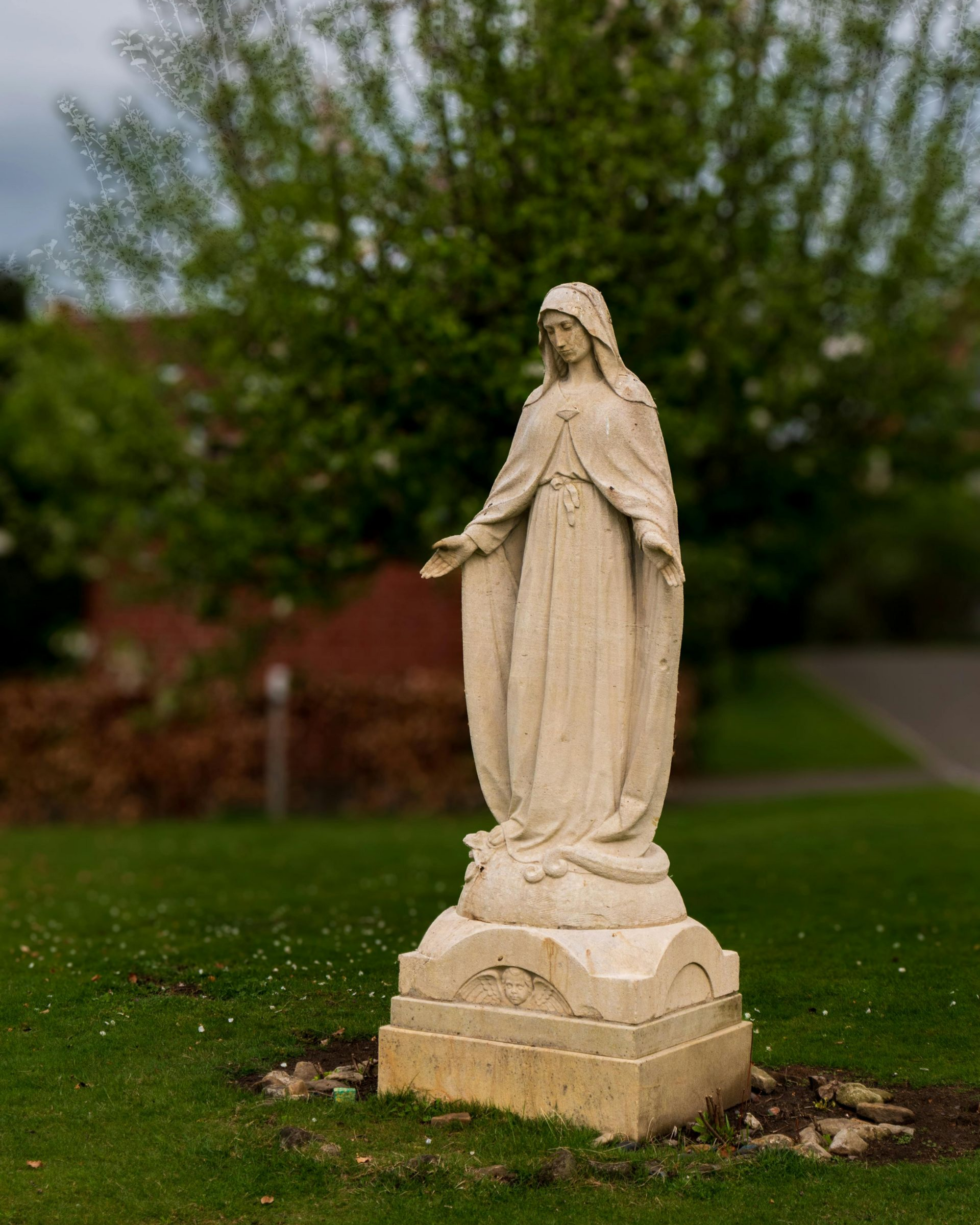 Statue of a Mary statue with outstretched hands on a stone pedestal in a grassy area with trees in the background.