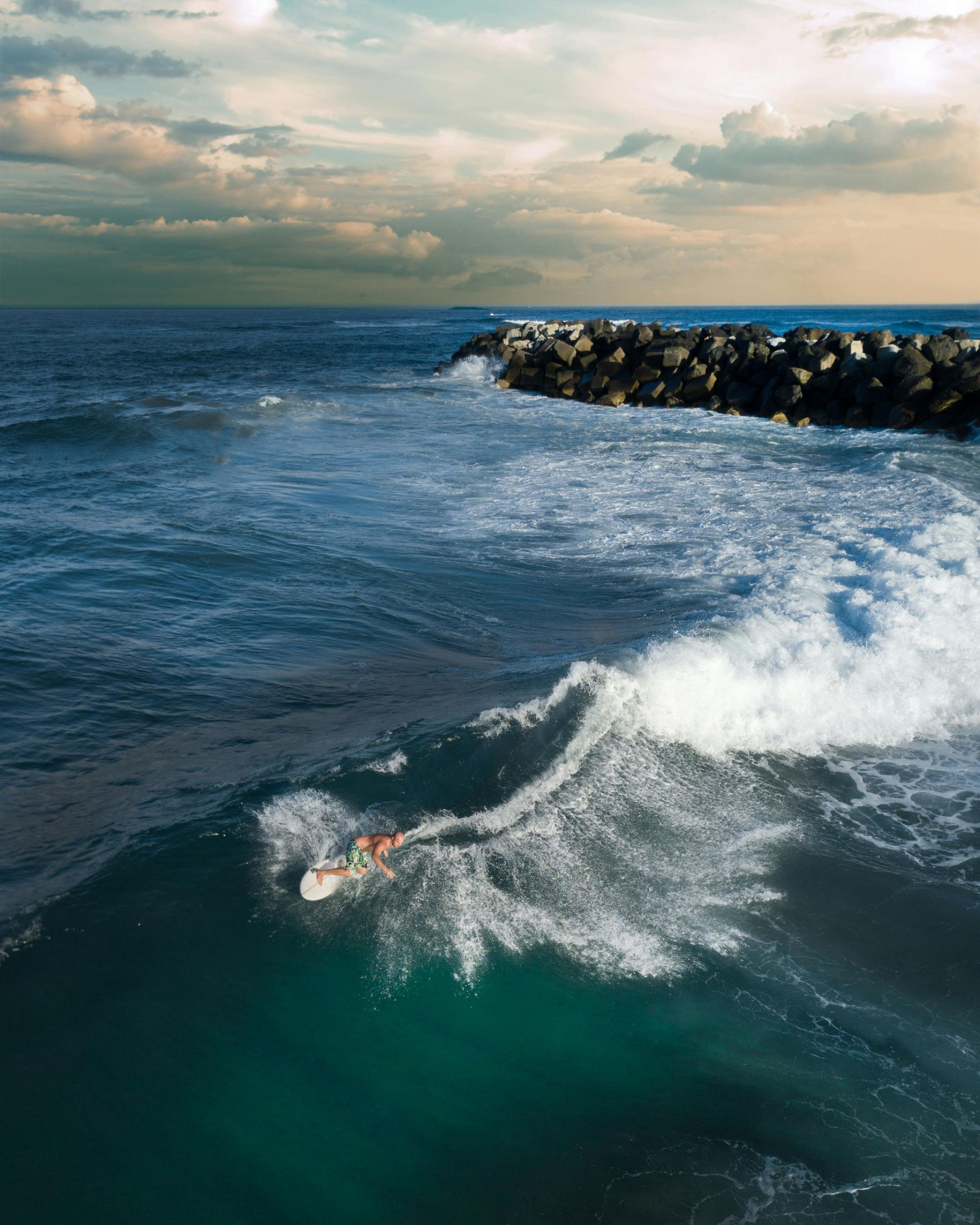 Surfer riding a wave near a rocky breakwater under a cloudy sky.