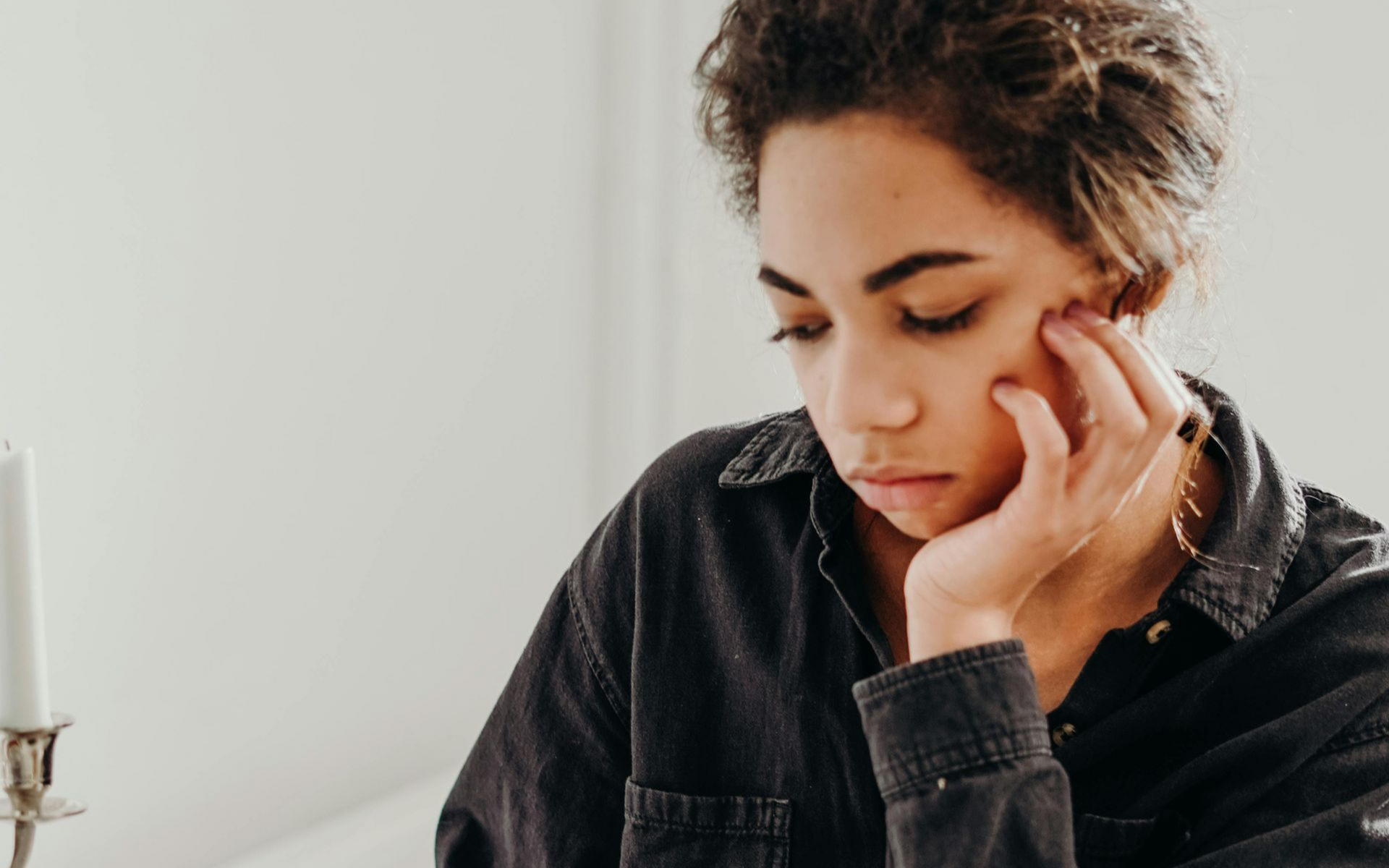 Woman with dark hair, resting chin on hand, looking down pensively. Interior, near a candle.