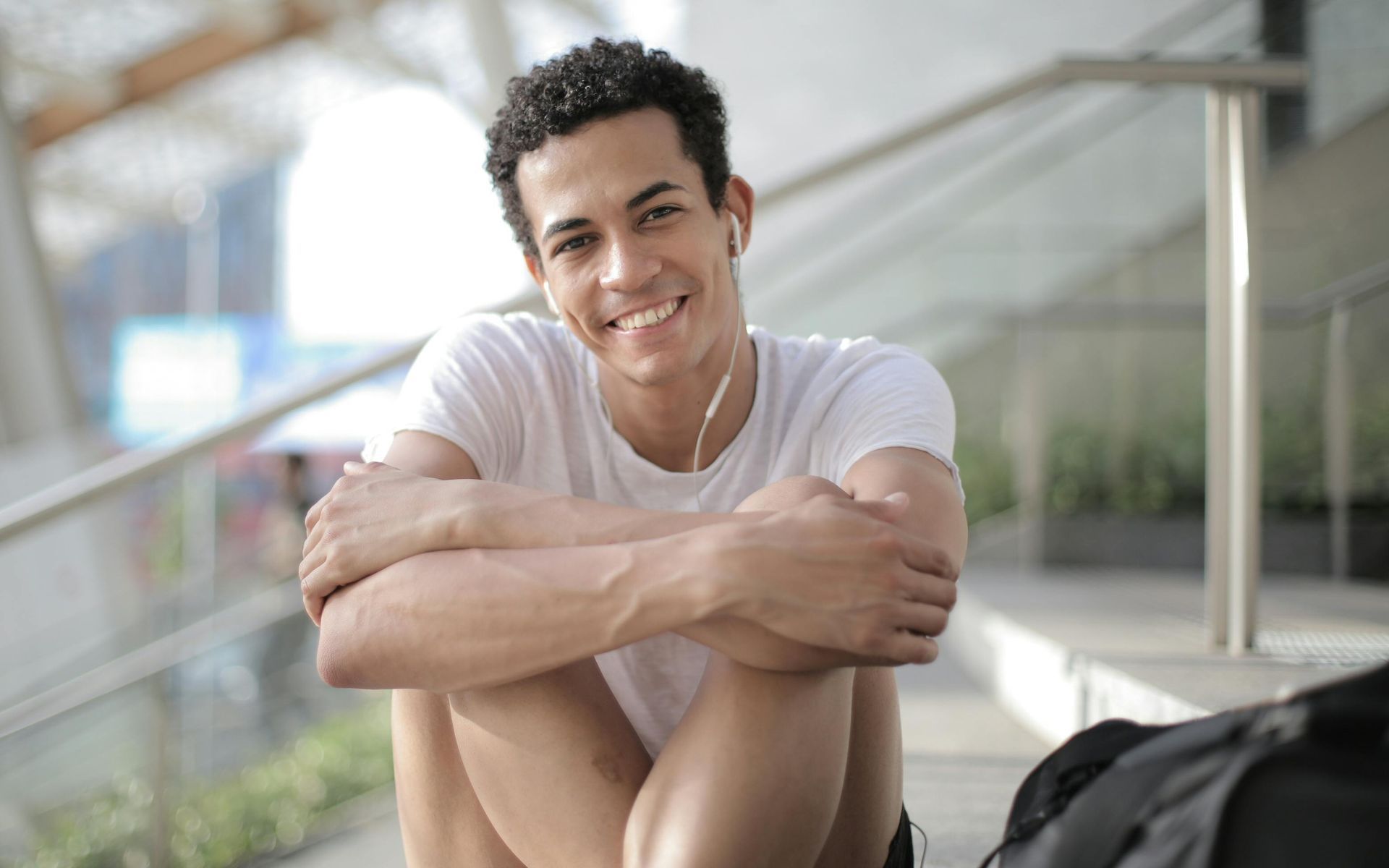Young person smiling, sitting on steps with arms crossed, earbuds in.