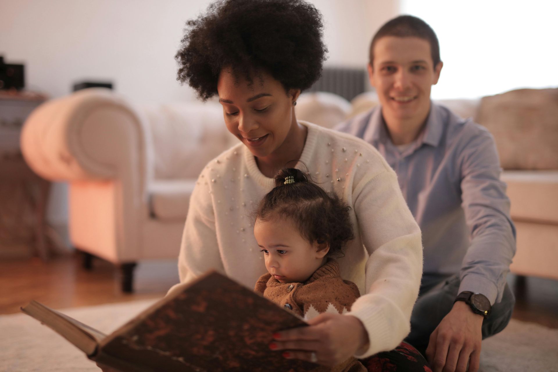 Woman and child read a book while man looks on, sitting near a sofa in a living room.