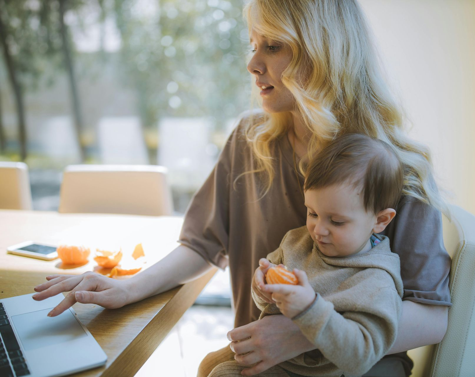 Woman working on laptop, holding a baby. Table has oranges and phone. Bright room with window.