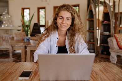 Woman with curly hair smiles while working on a laptop at a wooden table. A phone lies beside the laptop.