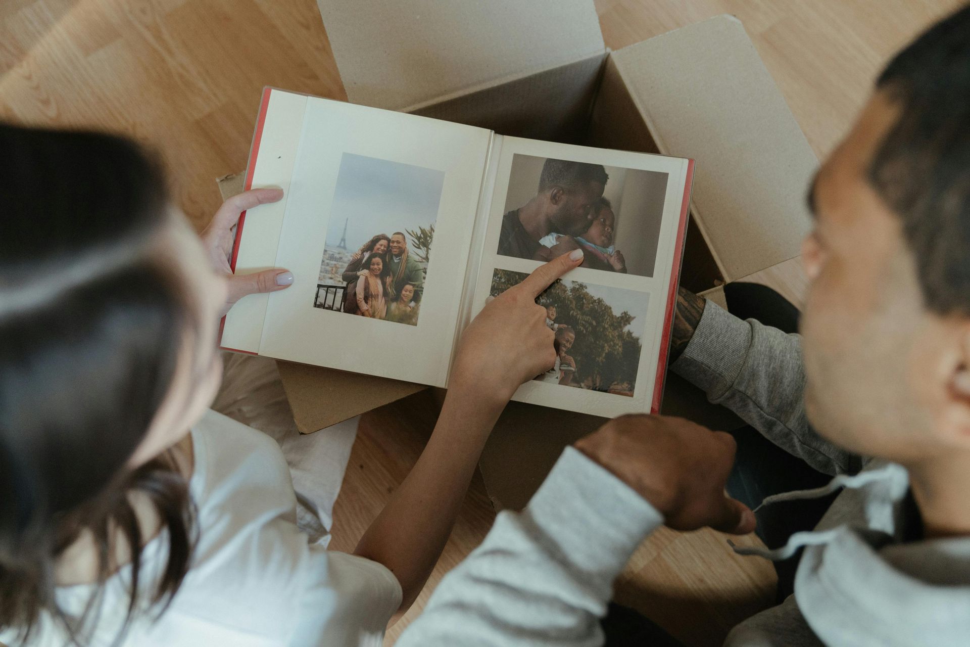 Couple looking at a photo album, pointing at pictures while sitting on a floor near a cardboard box.