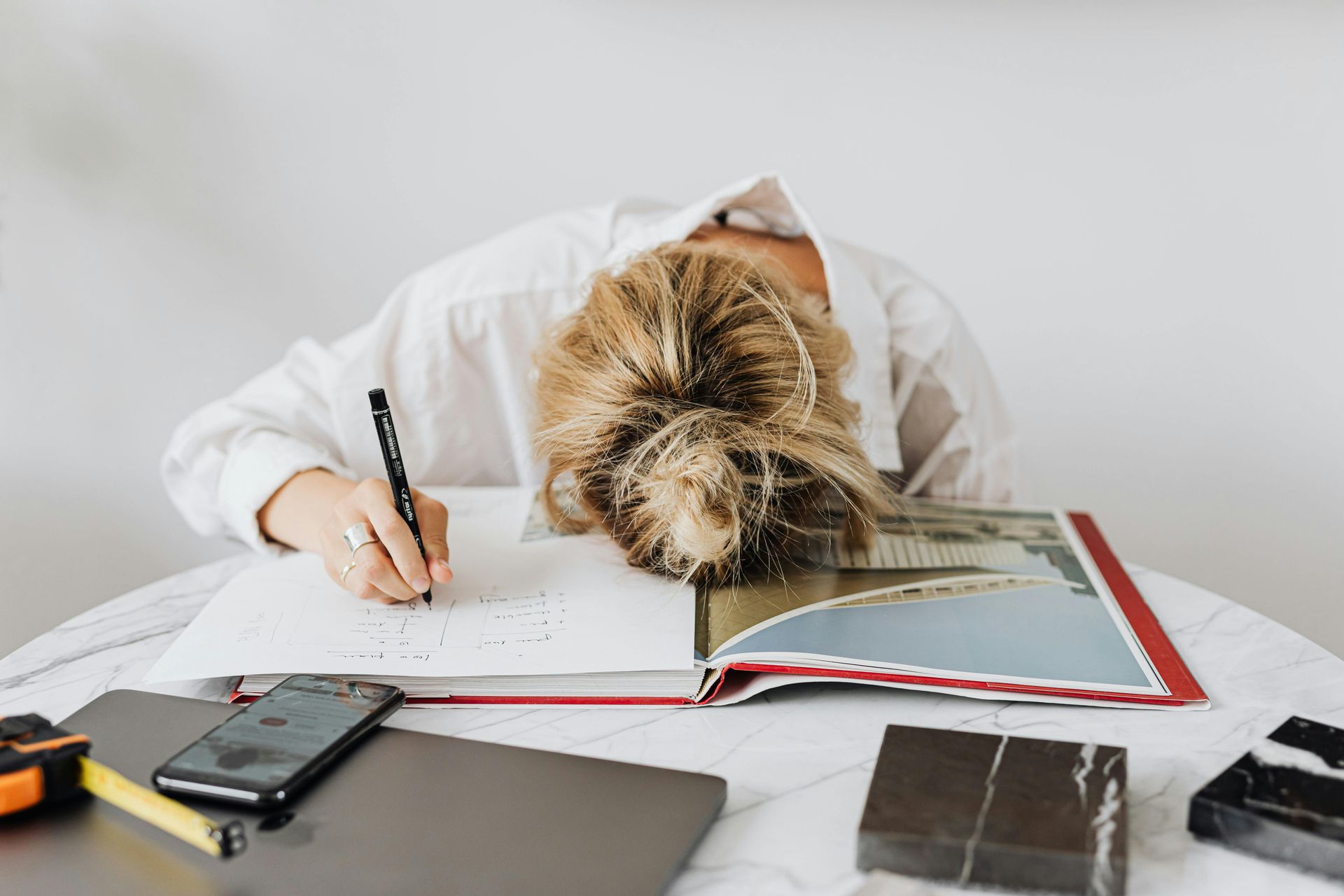 Person with head resting on desk writing in notebook. Scattered objects, workspace.