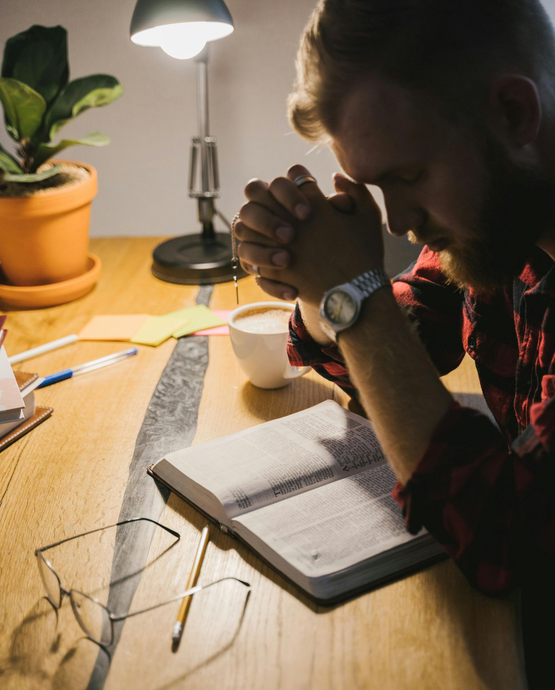Man praying with clasped hands over an open book, coffee, desk lamp, and potted plant.