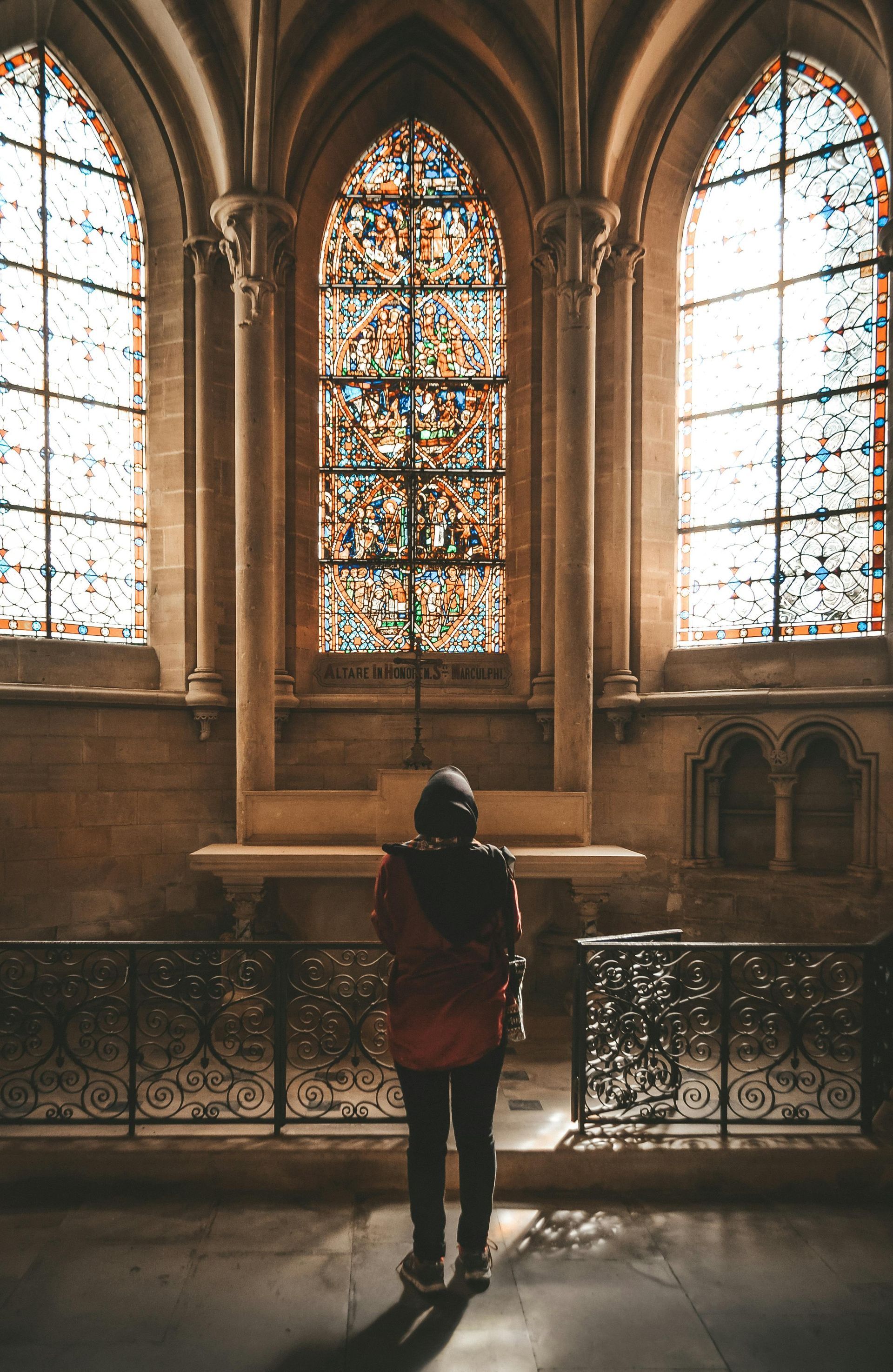 Person in a red jacket stands before a stained glass window inside a church.