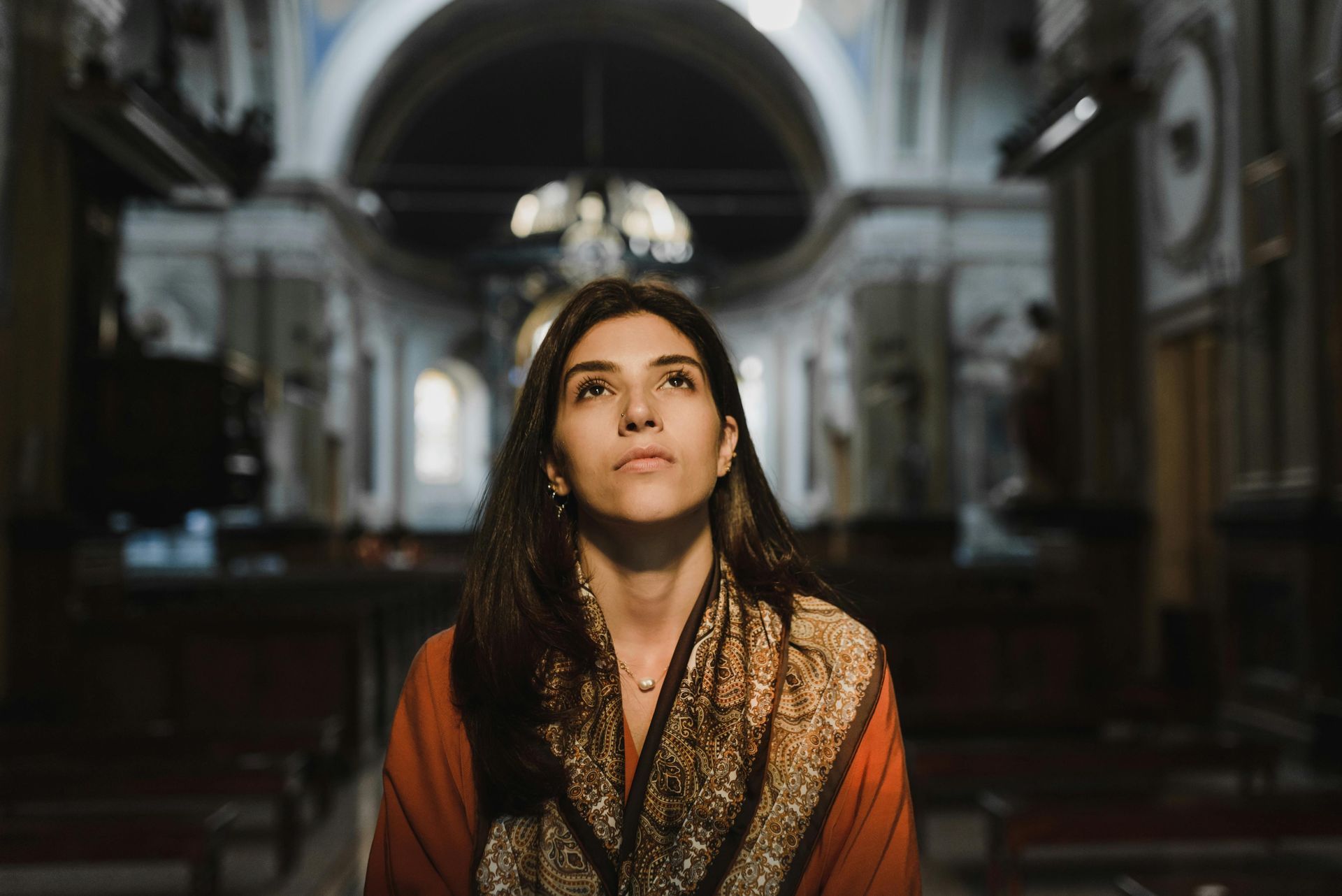Woman looking up in a church, wearing a patterned scarf, with a dark interior.