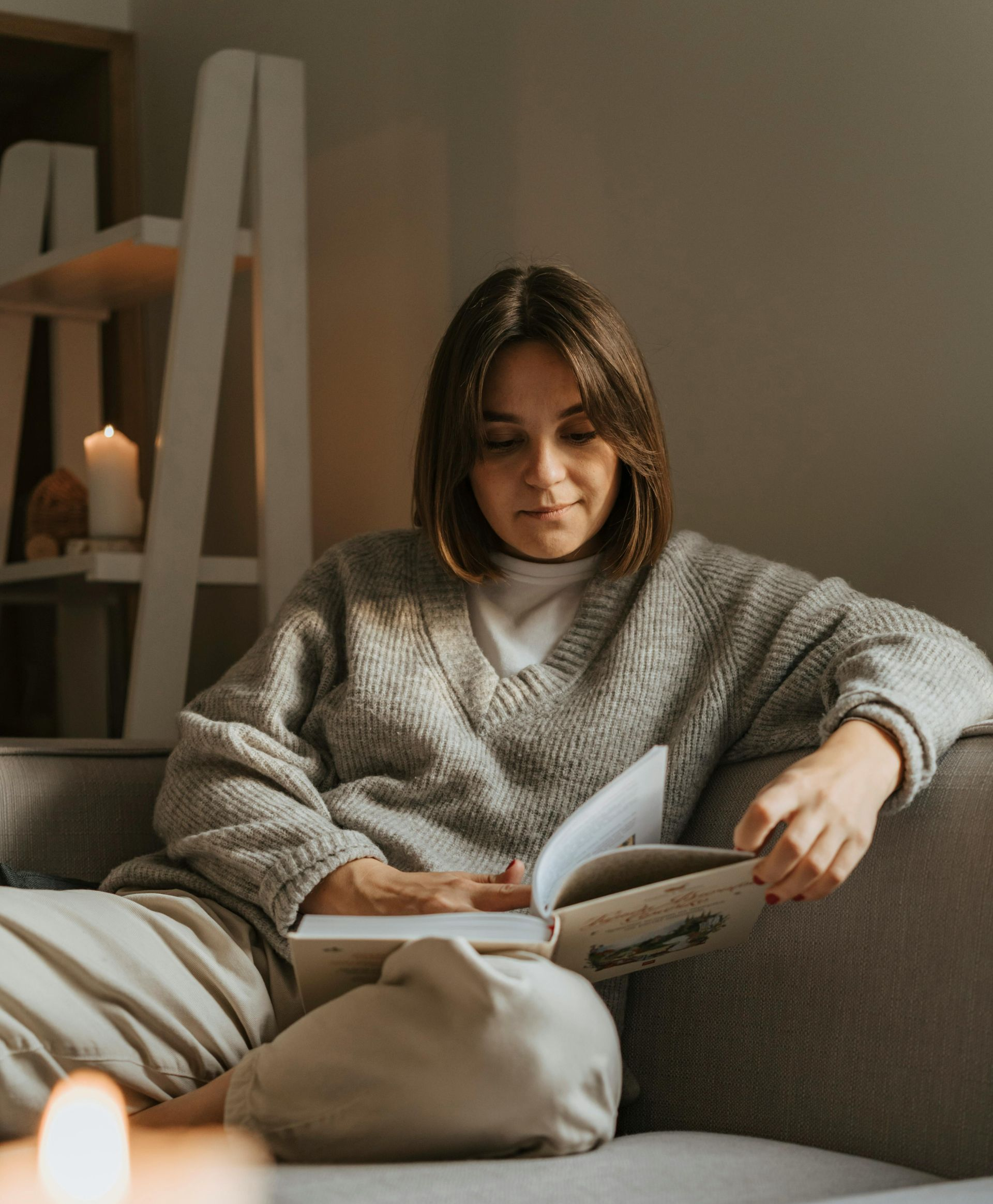 Woman in a gray sweater reads a book on a couch, lit by candles and a shelf in the background.