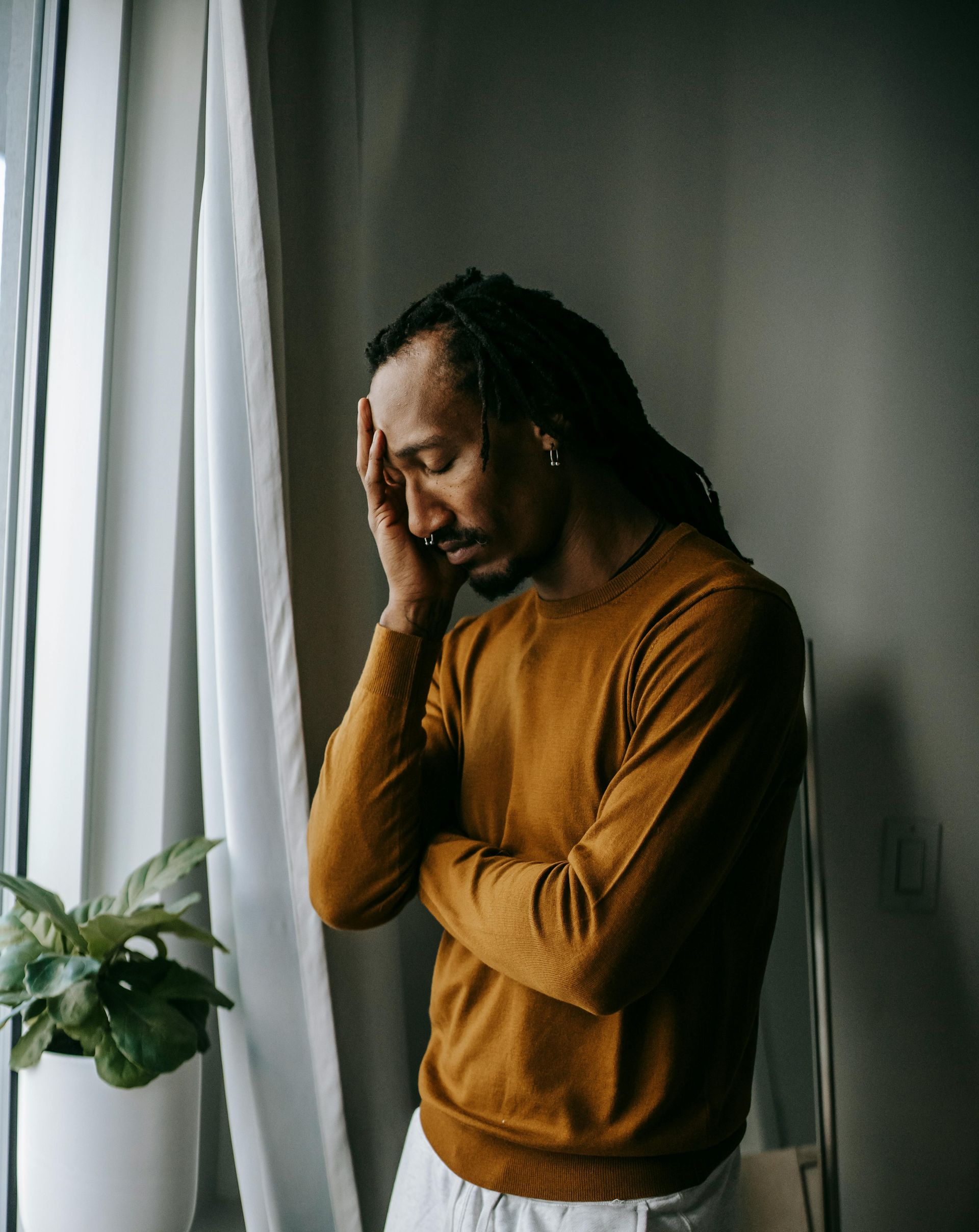 Man with dreadlocks, eyes closed, hand on forehead, standing near a window. He appears distressed.